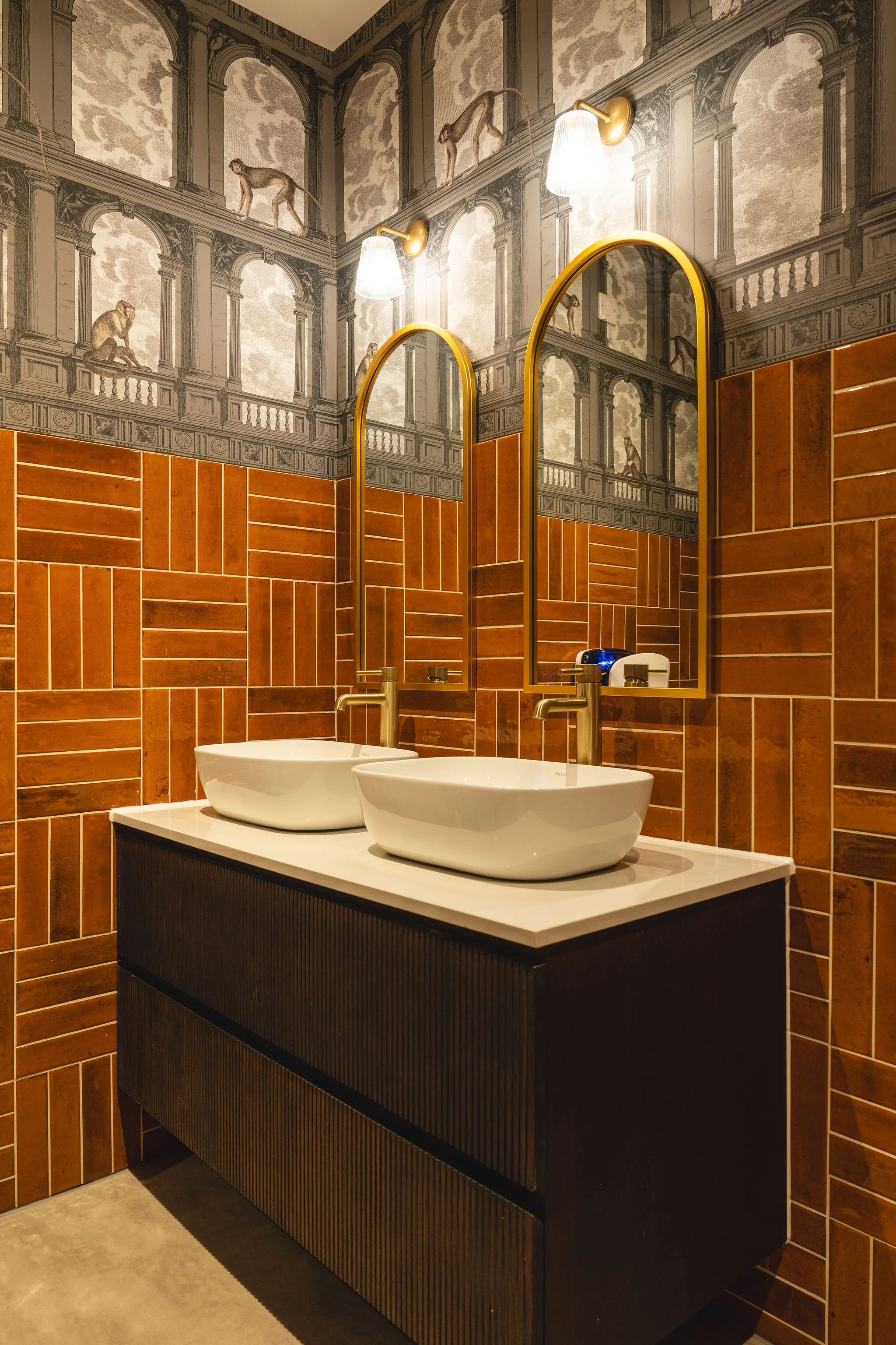 A bathroom with two white vessel sinks on a black vanity with vertical grooves. Behind the sinks, there are two large, gold-framed mirrors and two wall-mounted white light fixtures. The upper part of the wall features wallpaper with a mural resembling a classical architectural scene with animals like monkeys and giraffes.