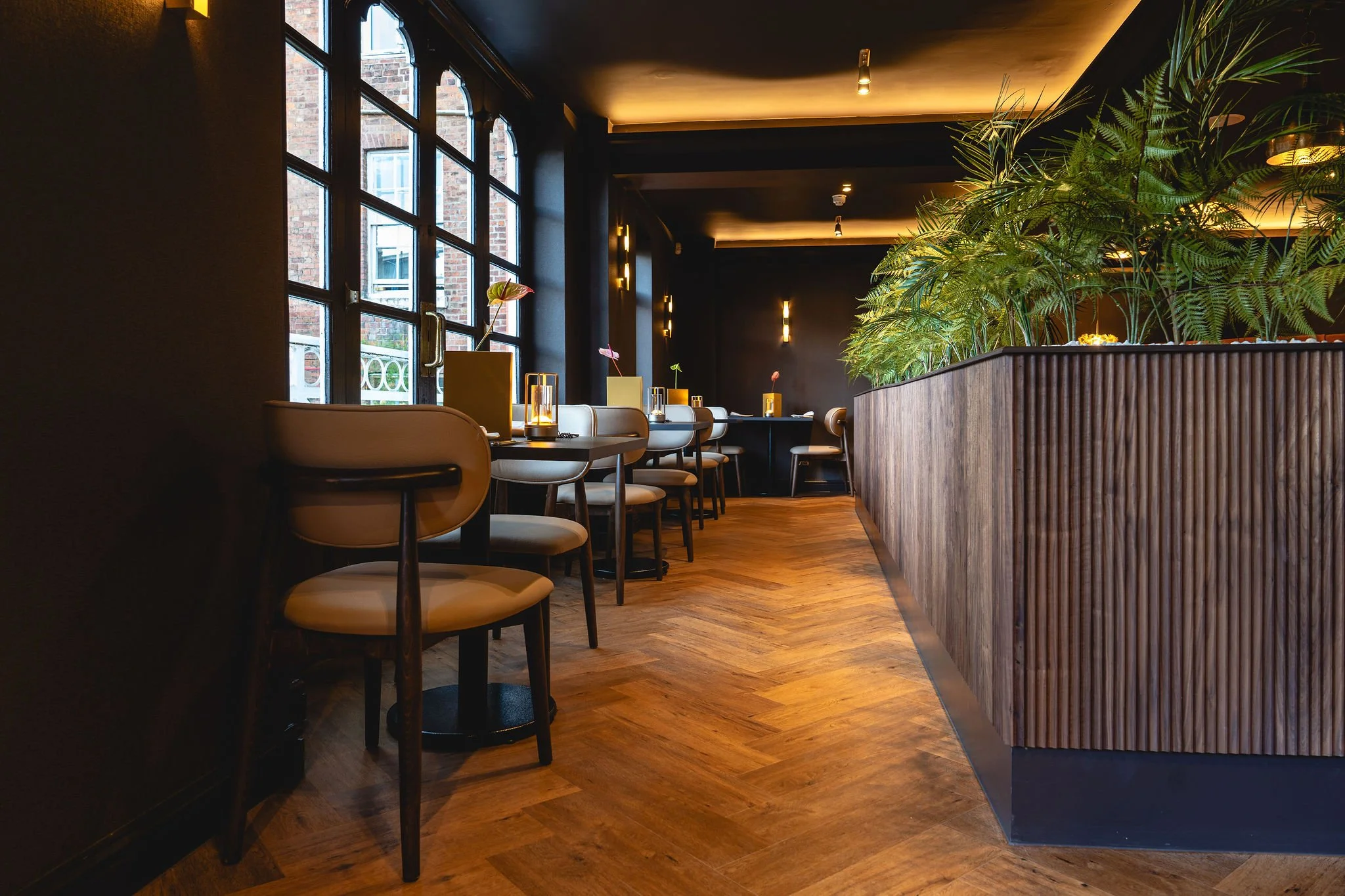 Interior of a modern restaurant or cafe with a row of tables along a large window, decorated with plants in yellow vases, and a wooden partition separating the space. Dim lighting creates a cozy atmosphere.