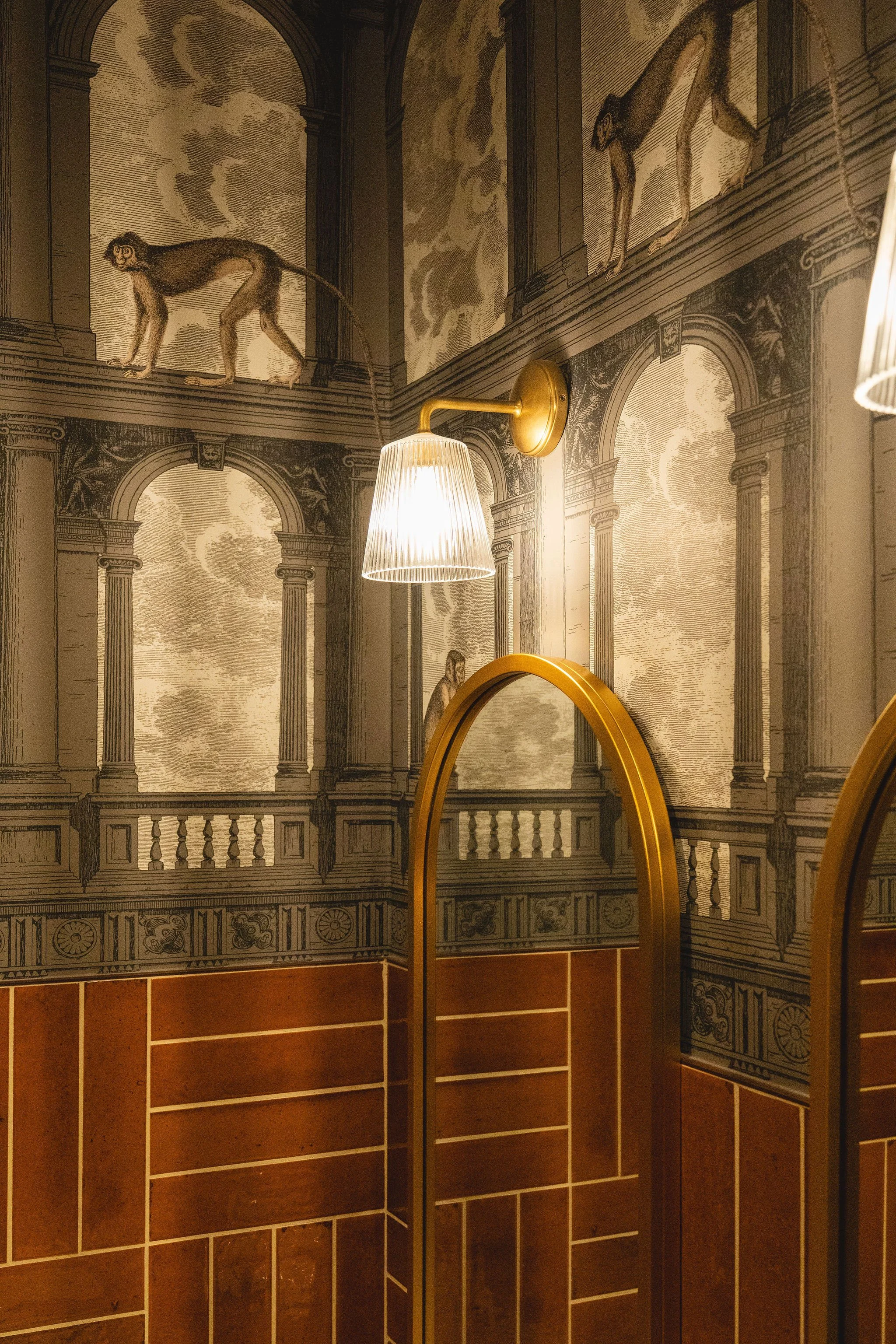 Interior of a room with walls covered in patterned wallpaper depicting classical architectural elements and monkeys. A rectangular mirror with a gold frame is reflected in the picture. There is a brass wall-mounted lamp with a pleated lampshade illuminating the space. At the bottom, there are shelves or storage compartments with a reddish-brown finish.