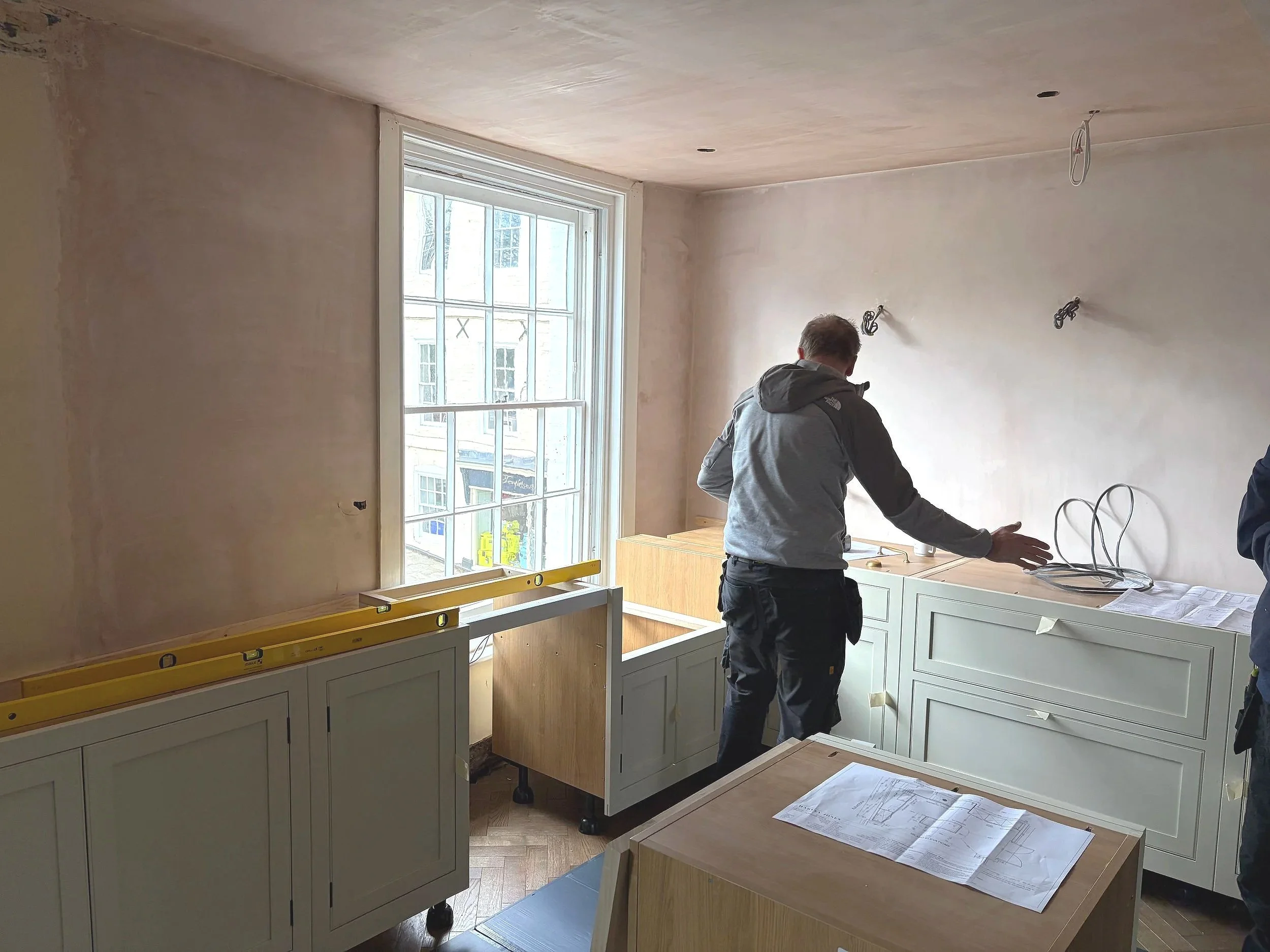 Interior of a room under renovation with a man working, cabinets being installed, window allowing natural light, construction tools, and plans on a table.