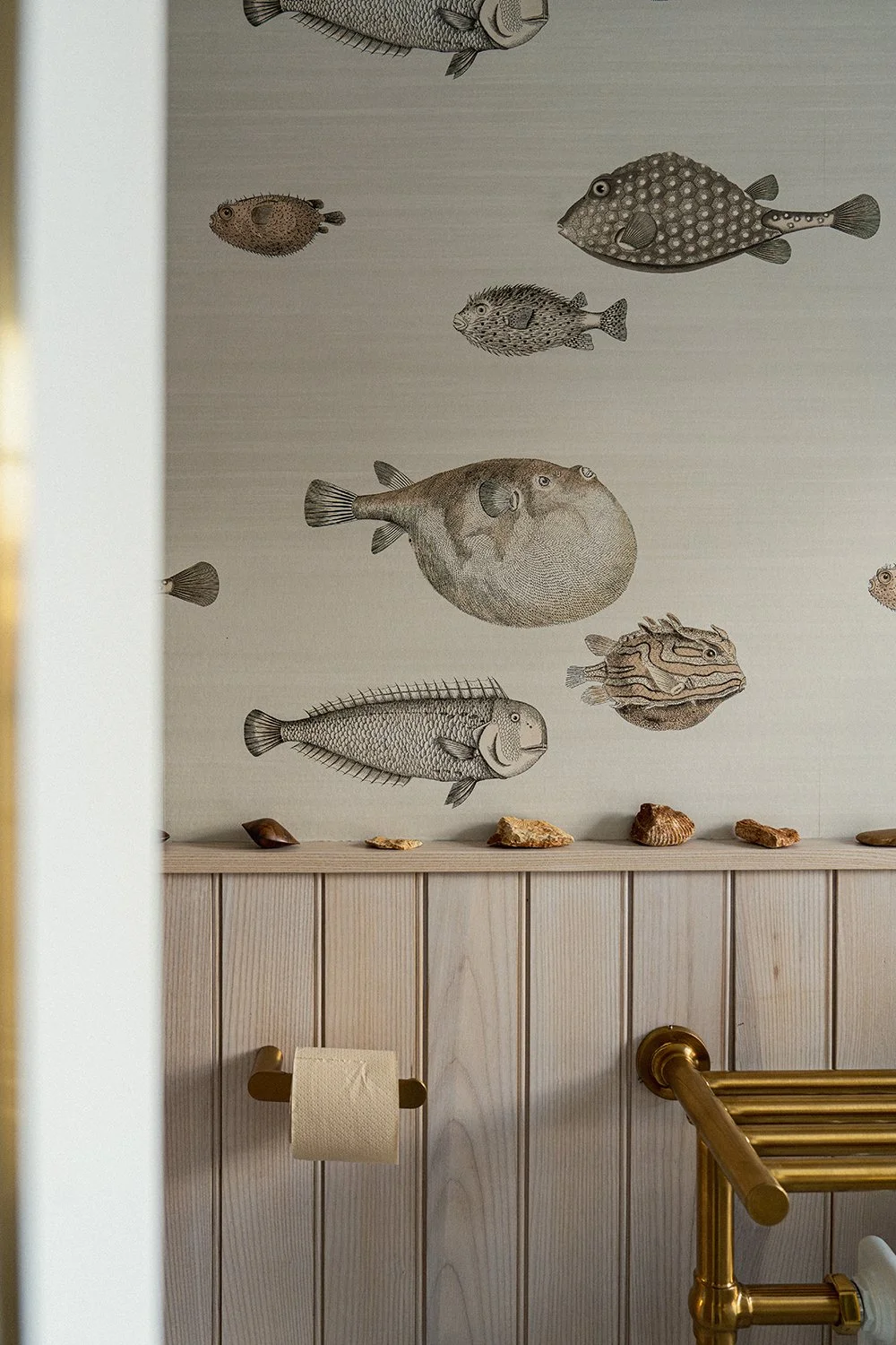 Contemporary shower room featuring fish-print wallpaper, brass toilet roll holder, timber panelling, and collected coastal-inspired styling.