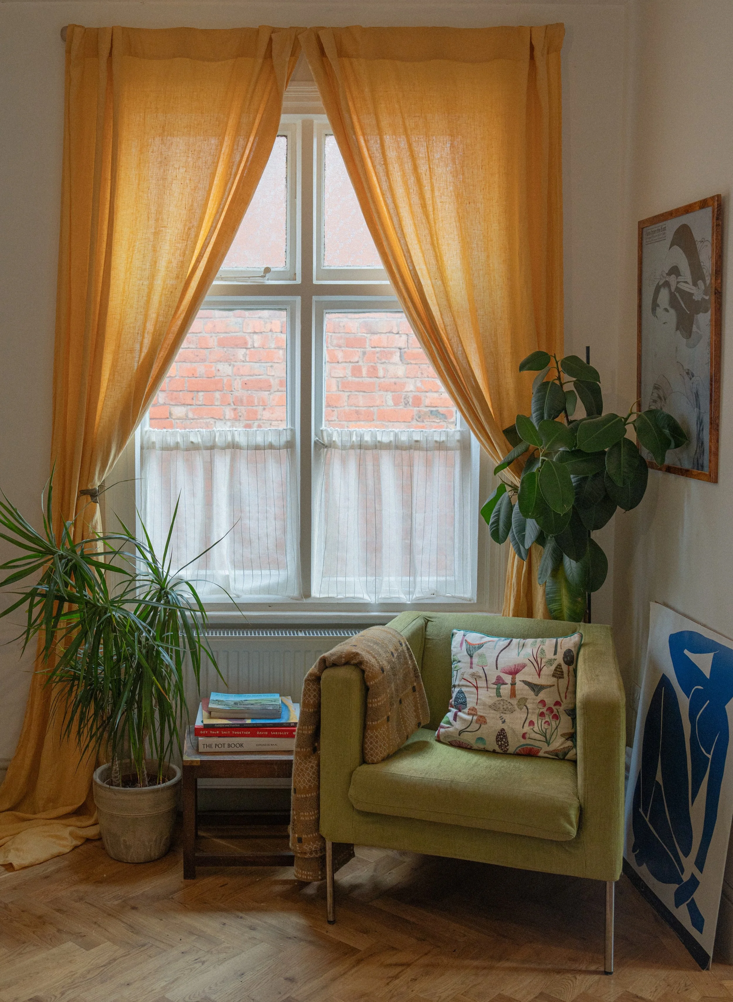 Living room corner with large window and yellow curtains. A green armchair with a colorful mushroom-patterned pillow, a small side table with books, and a potted plant are visible. Framed art on the wall and parquet flooring complete the scene.