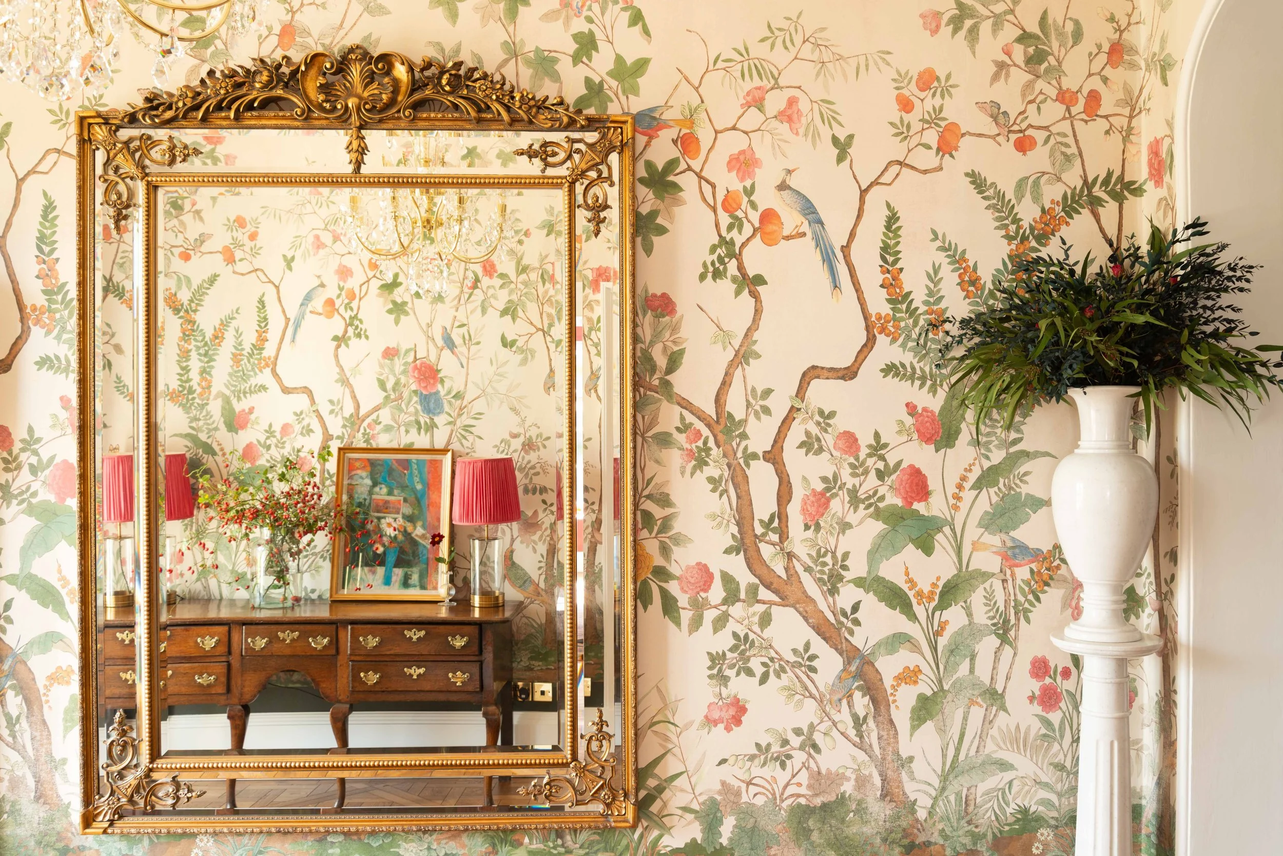 A decorative interior room with floral wallpaper, a large ornate gold-framed mirror, a wooden dresser, framed artwork, red lampshades, a vase with flowers, and a white pedestal with a large plant.