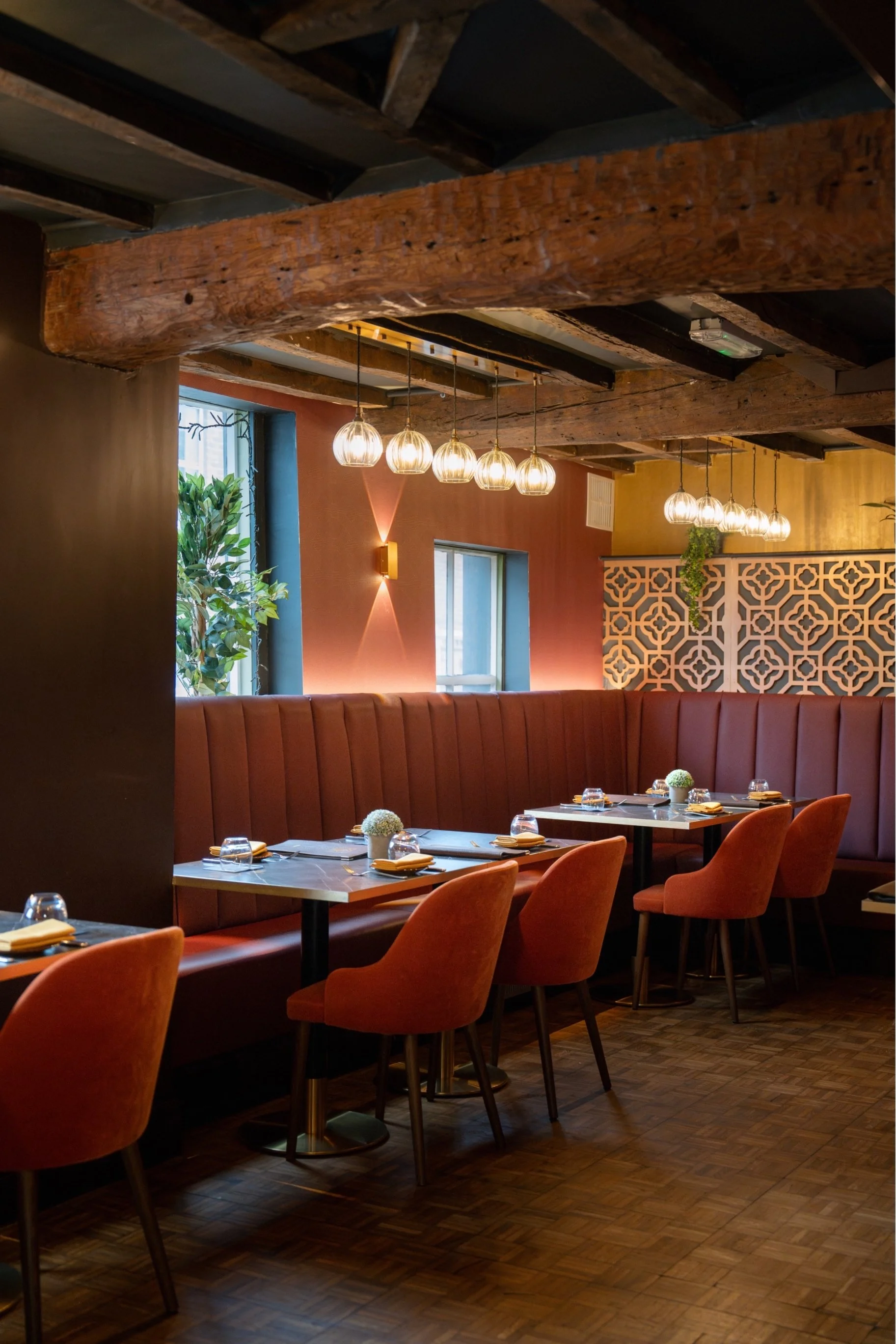 A cozy, stylish restaurant interior featuring a wooden beam ceiling, pendant lighting, and a long curved booth with orange chairs around square tables set with glassware and napkins. There are large windows with natural light and lush green plants.
