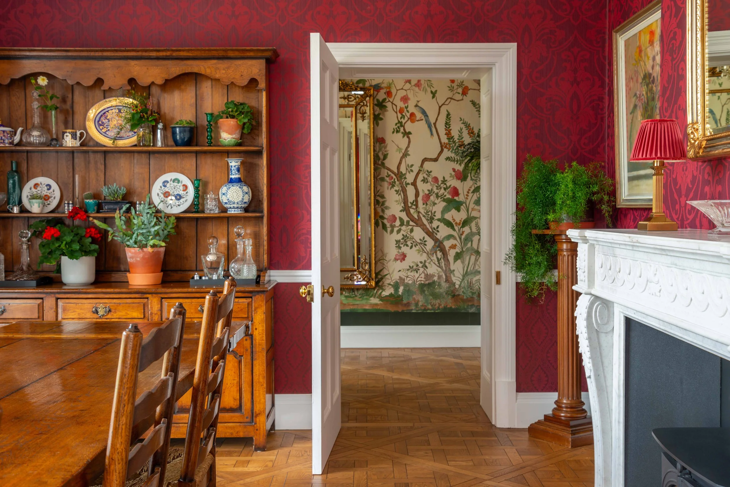 Richly layered Georgian dining room with burgundy wallpaper, antique wooden furniture, ornate fireplace, and botanical mural creating a dramatic interior scheme.