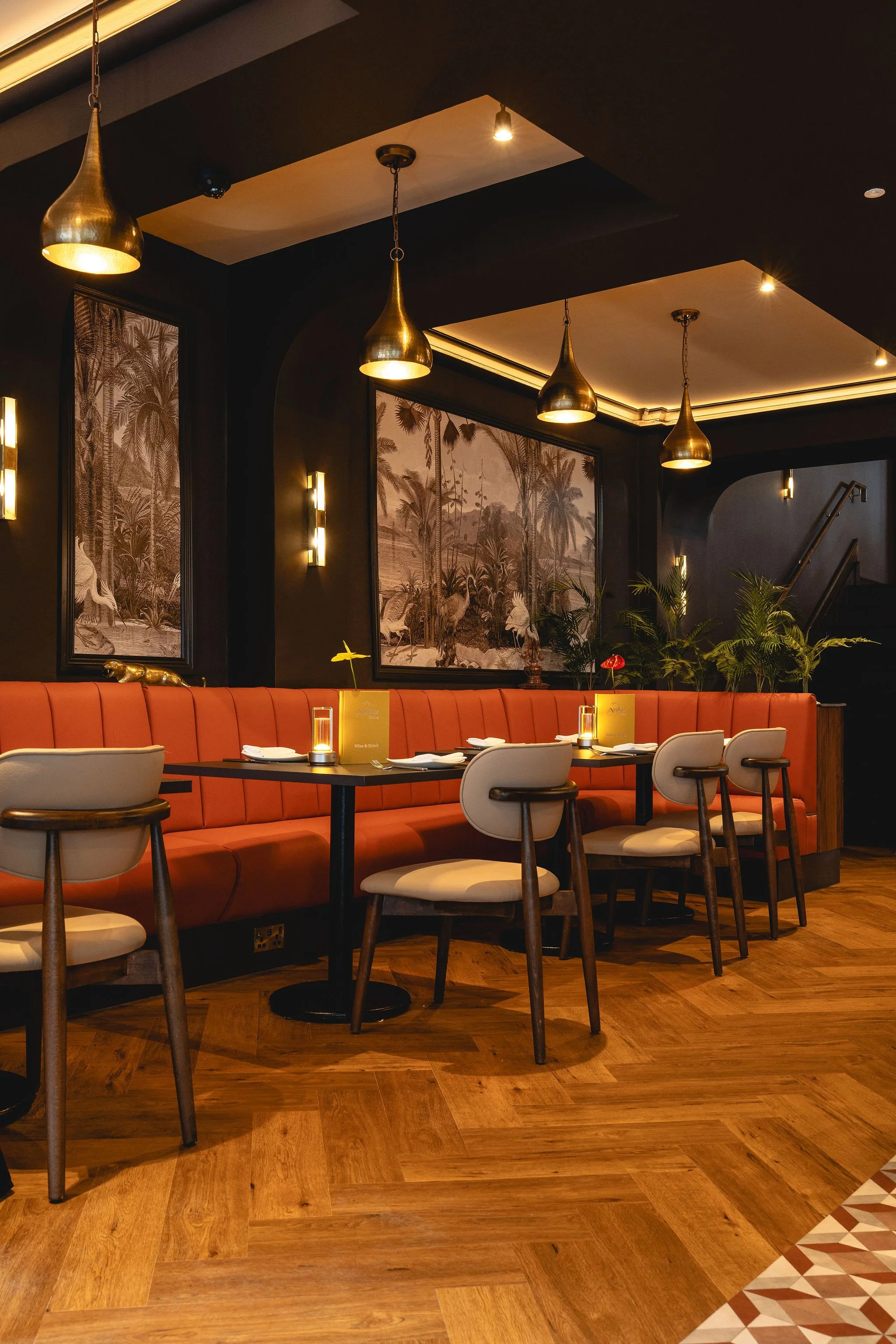 Interior of a stylish restaurant with a long red banquette, wooden chairs, round tables set with white plates and small candles, framed black-and-white tropical landscape artwork on dark walls, gold pendant lighting, and warm lighting accents.
