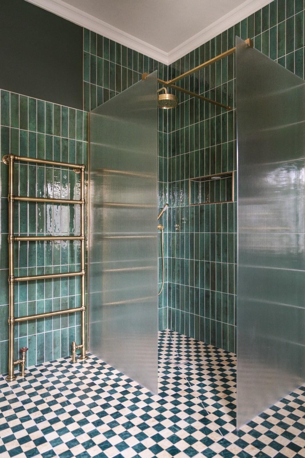 Modern shower with green tiles, a brass towel radiator, a glass shower partition, a shower head, and black-and-white checkered floor tiles.