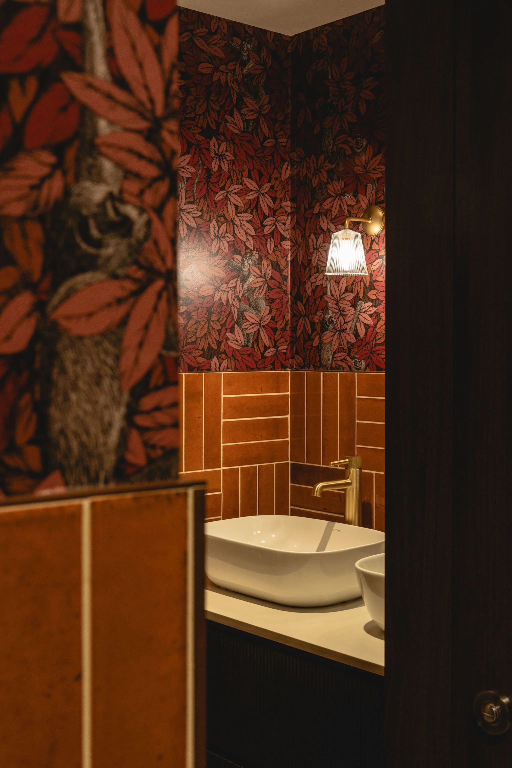 A bathroom with floral wallpaper featuring reddish leaves and trees with faces, a modern gold sconce with a glass shade, a white oval vessel sink, and a smaller white bowl next to it.