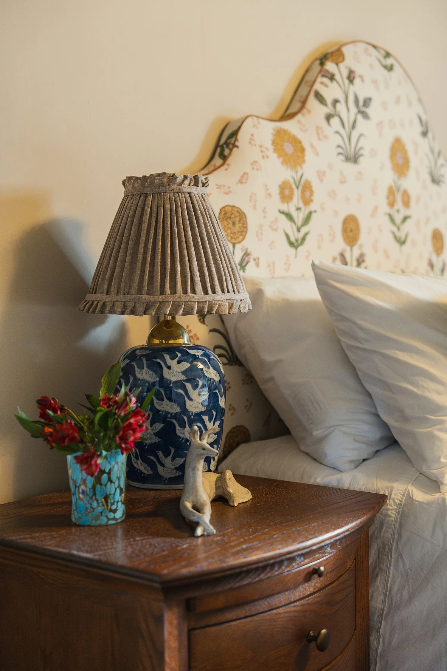 Styled bedside vignette with pleated lamp shade, floral headboard, antique furniture, and fresh flowers in a welcoming guest bedroom.
