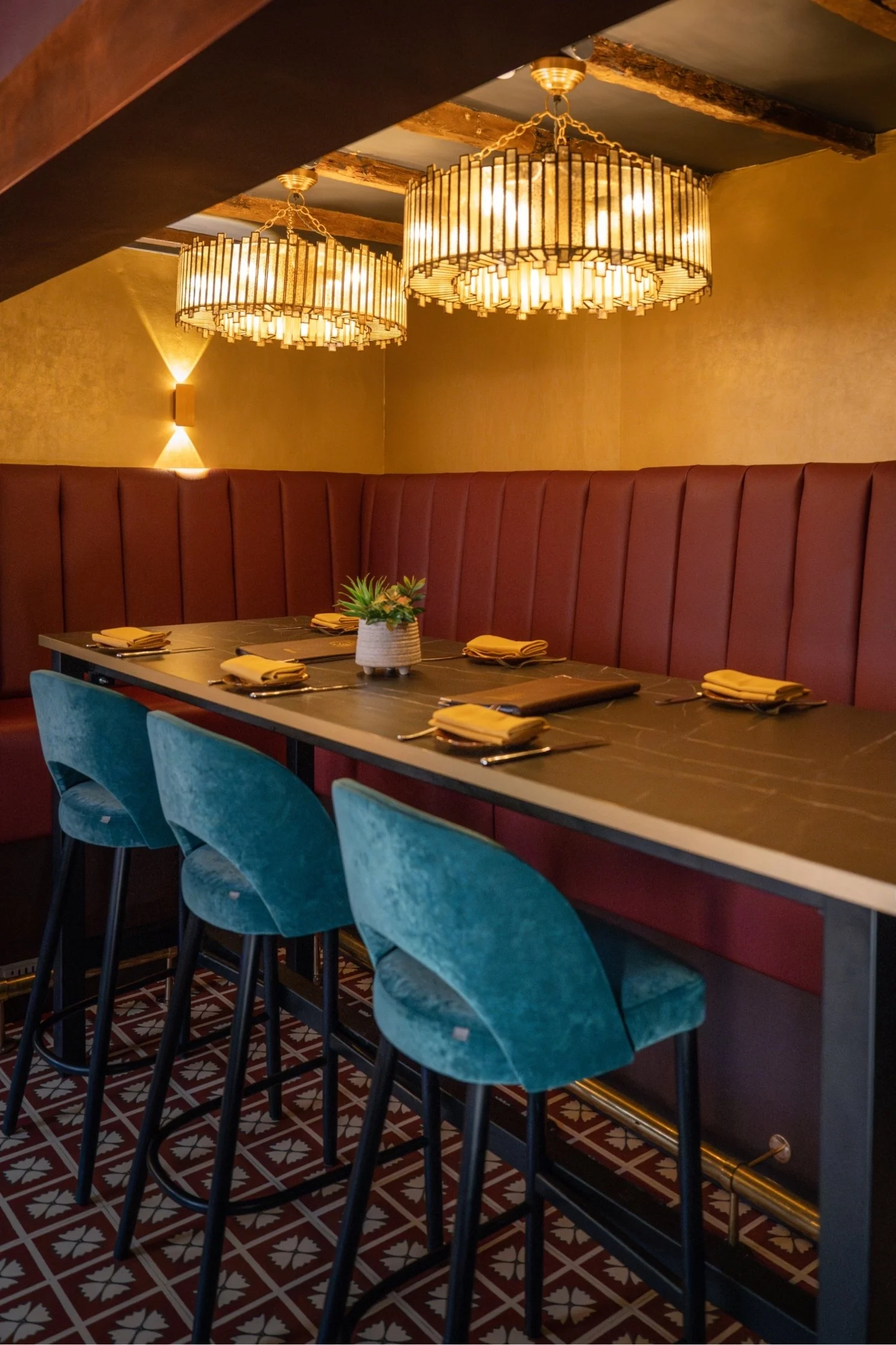 A restaurant dining area with a long table set with yellow napkins, blue chairs, a potted plant, and warm lighting from two chandeliers hanging from the ceiling. The background features a beige wall and patterned flooring.
