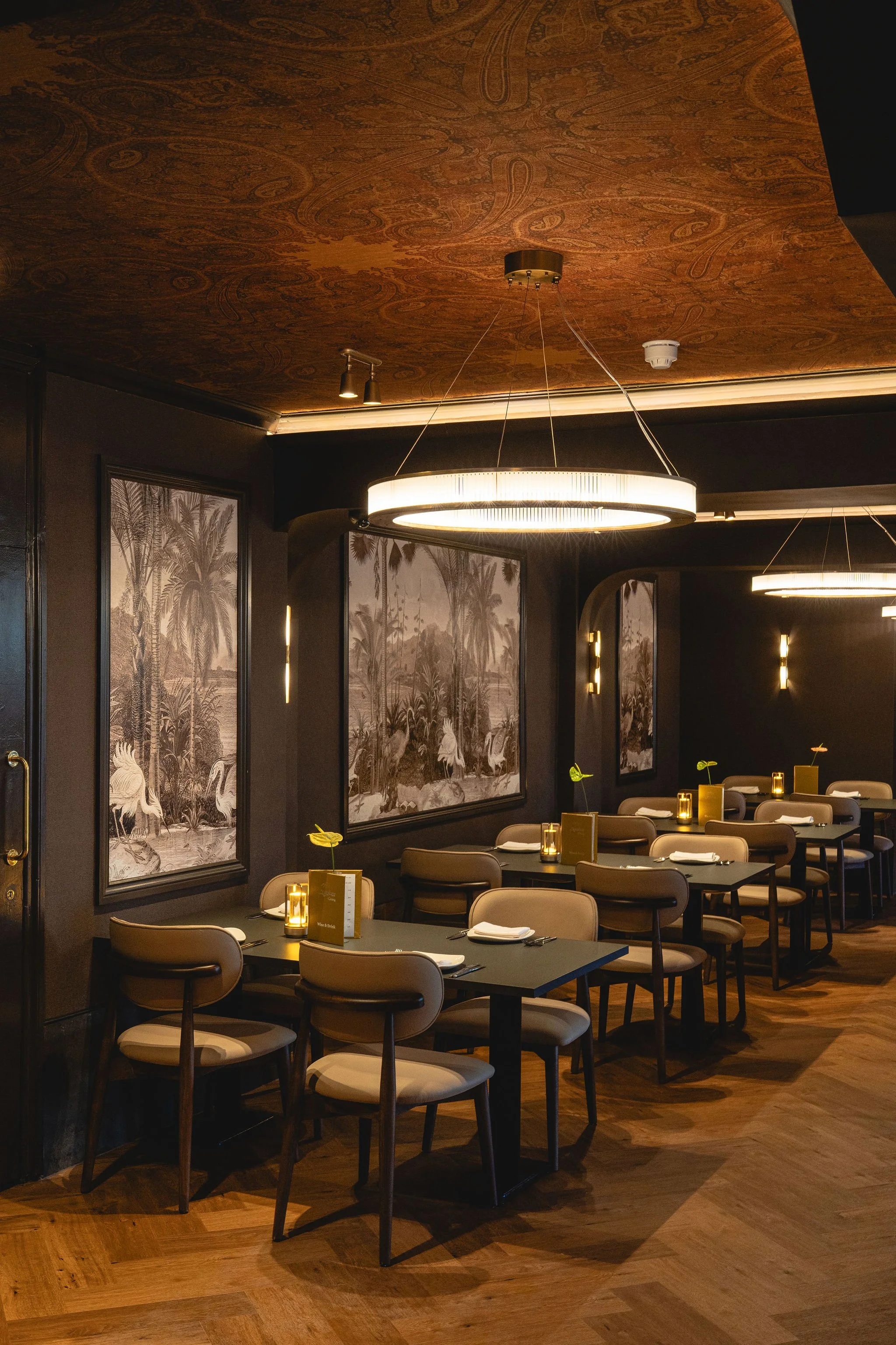A dimly lit restaurant interior with dark walls and wooden floors. Several tables with beige chairs are set with napkins, cutlery, and small table lamps. Framed black-and-white nature scenes hang on the walls. Modern circular pendant lights hang from the ceiling, providing warm lighting.