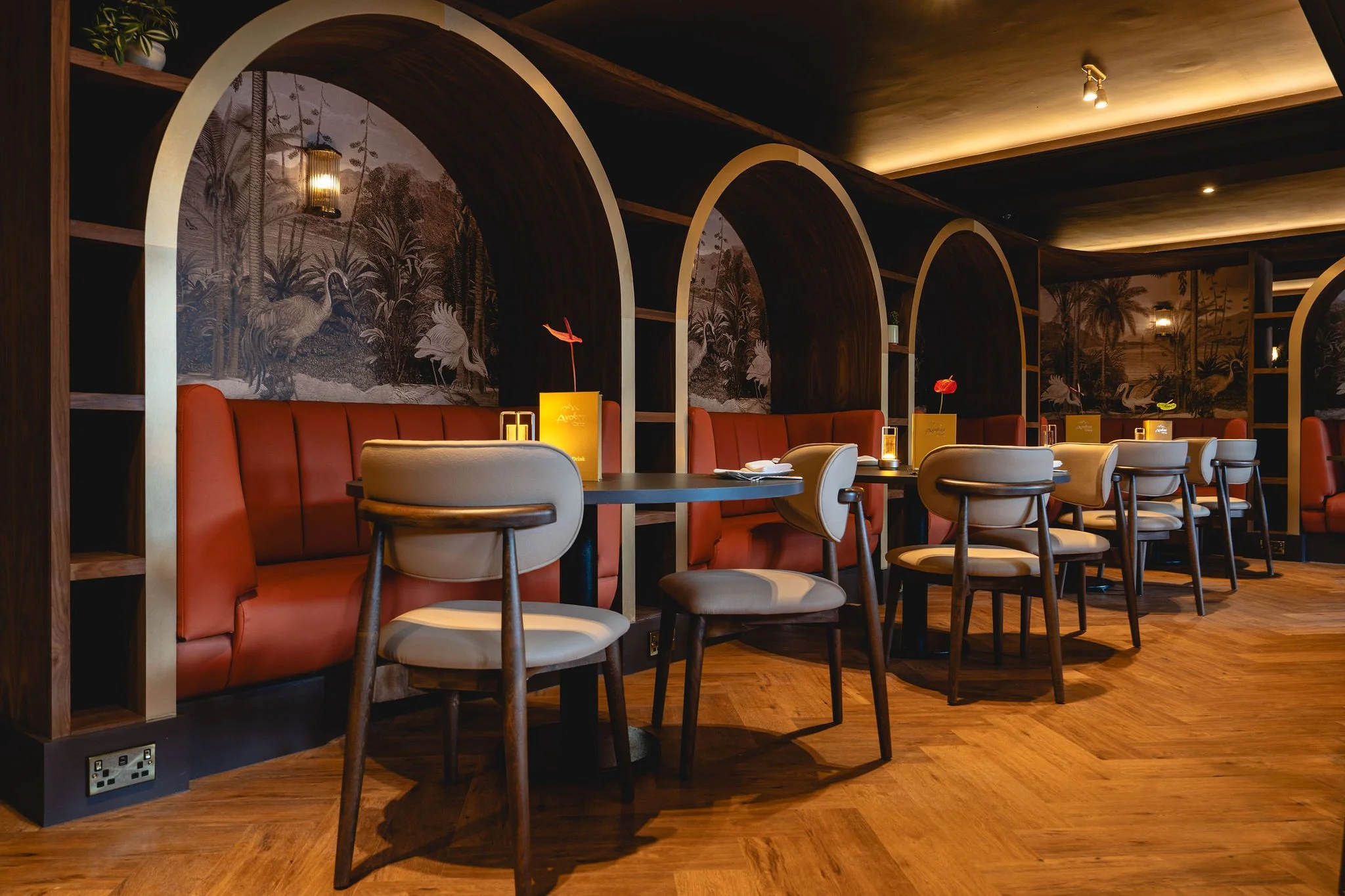 A restaurant interior features tables with white and brown chairs, a red booth, illuminated table lamps with flowers, decorative backdrops with wildlife murals, wooden shelving with potted plants, and warm lighting on a wooden floor.