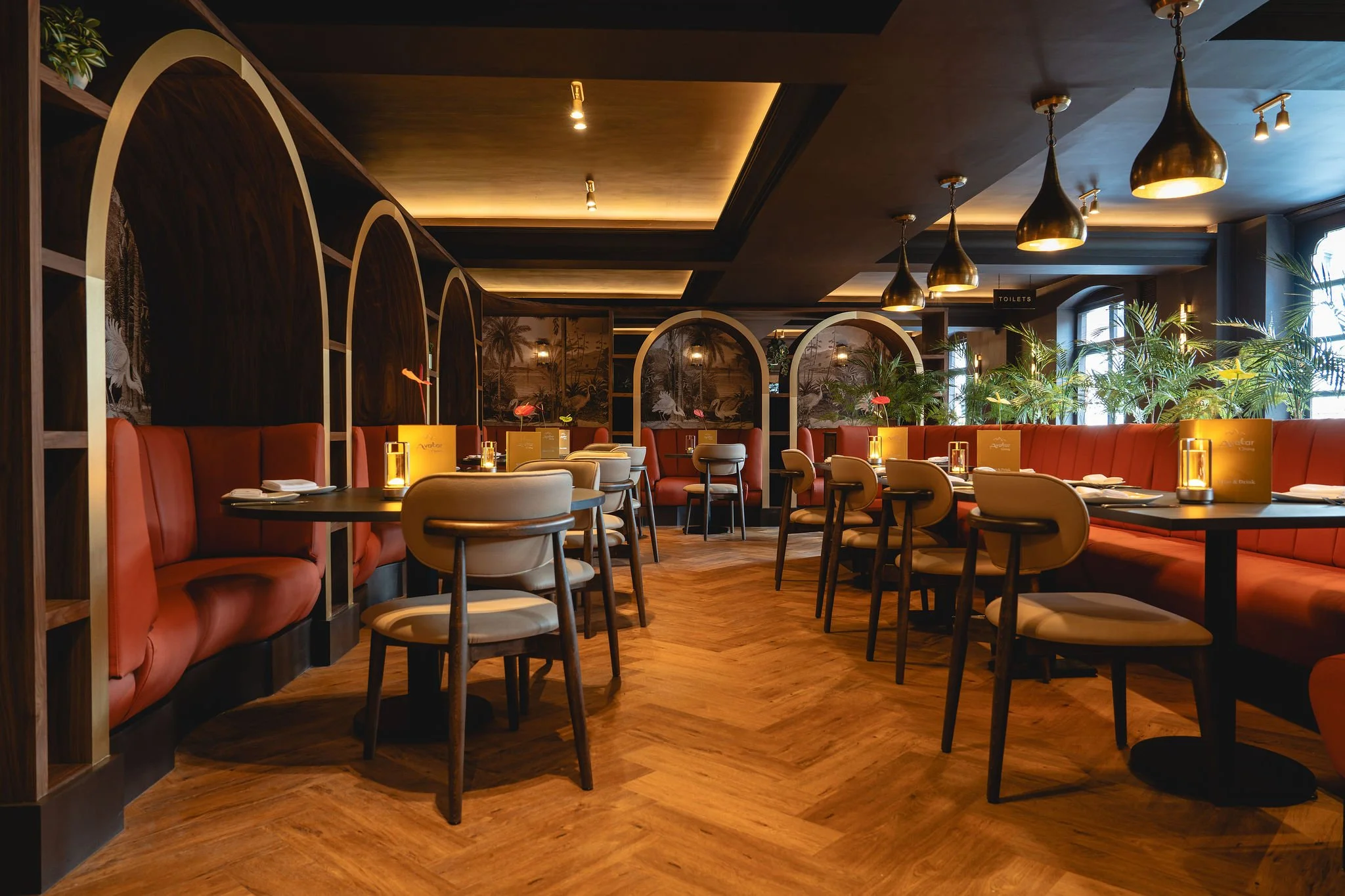 Interior of a modern restaurant with warm lighting, wooden floors, red and beige seating, decorative wall art of tropical scenes, and lush green plants near windows.