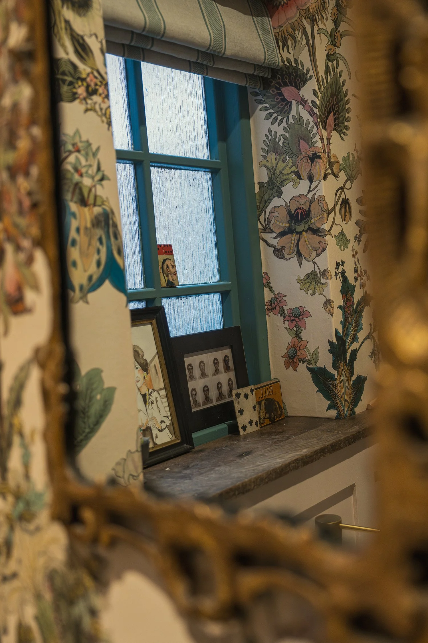 Decorative powder room detail with botanical wallpaper, blue-painted sash window, and antique brass mirror in a richly layered interior scheme.