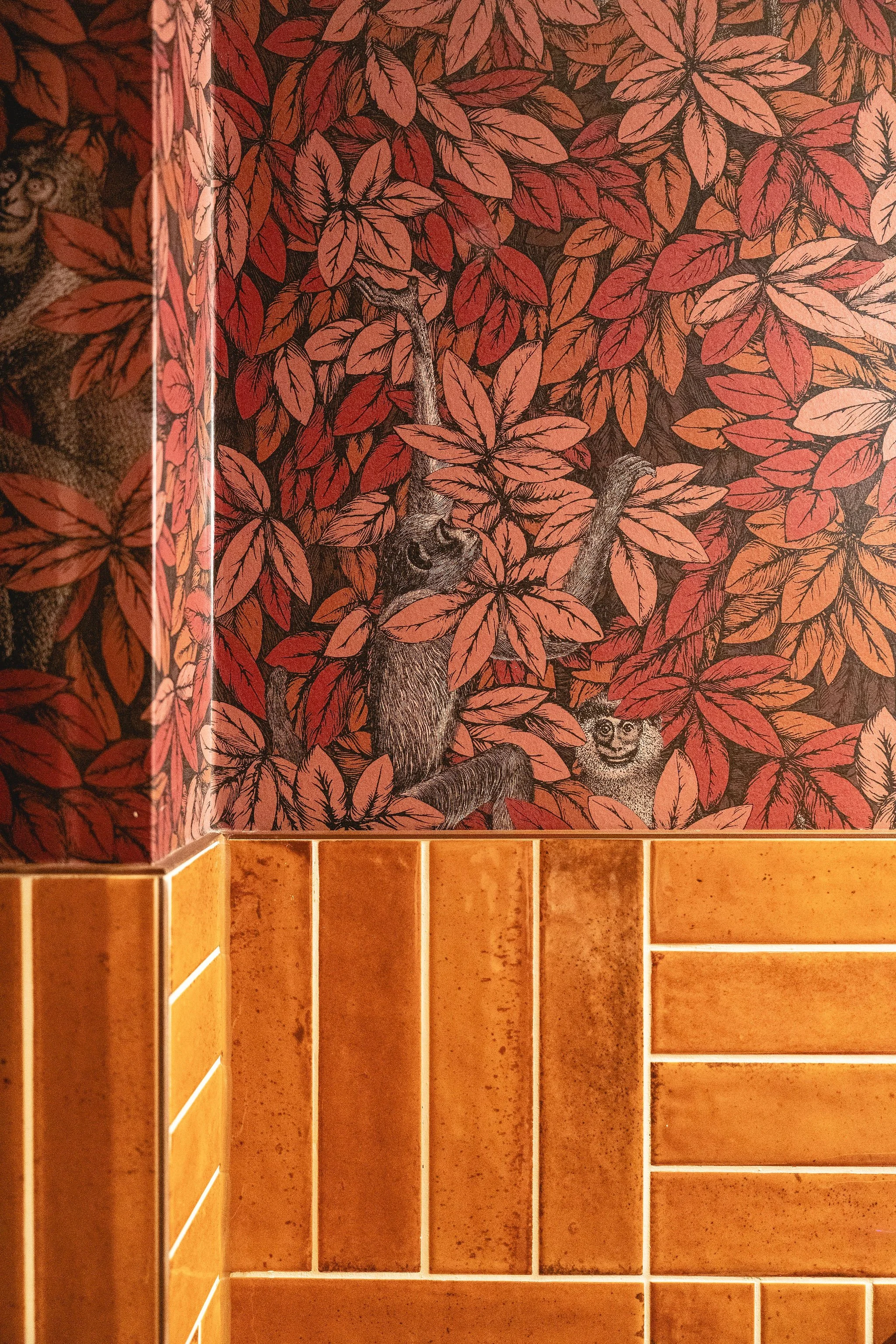 The image shows the corner of a room with a decorative wall covered in a leafy pattern featuring reddish and orange leaves, and illustrations of small foxes camouflaged among the leaves. The lower part of the image displays wooden cabinets or shelves with a warm, orange-brown finish.