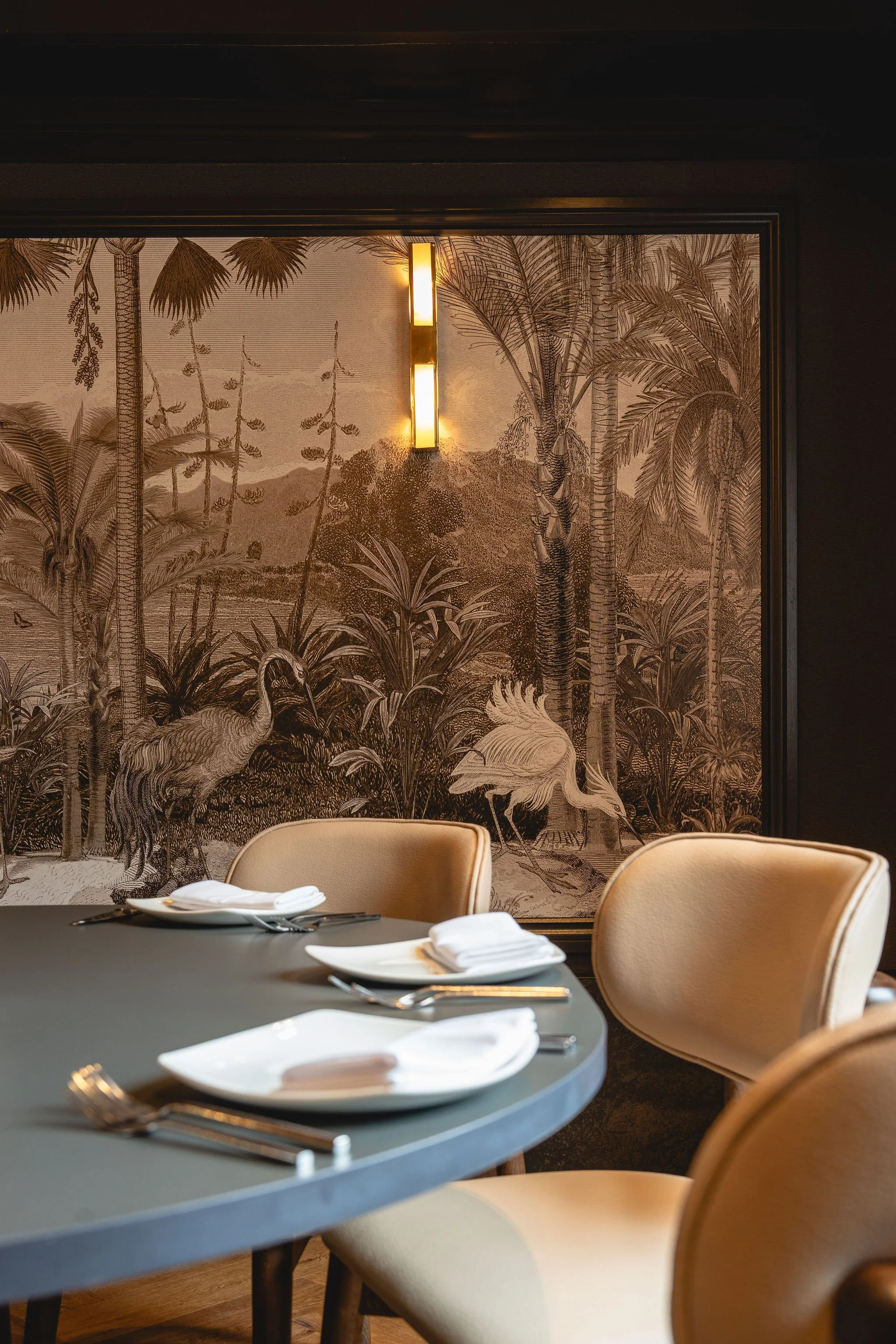 Interior dining room with a dark table set with white napkins and silverware, beige chairs, and a large tropical-themed mural featuring palm trees, foliage, and flamingos, with a modern wall-mounted light fixture.