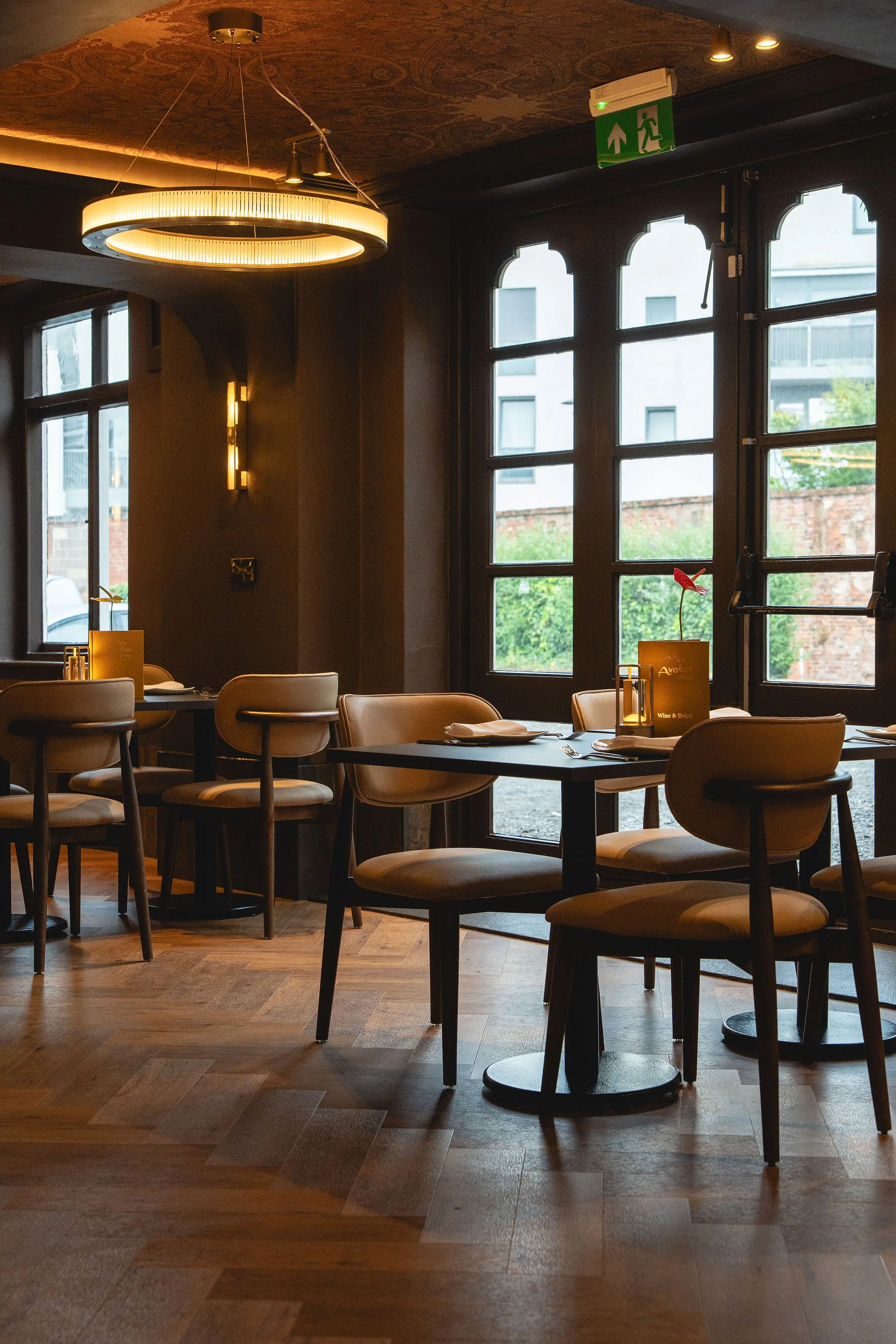 Interior of a restaurant or cafe with tables and chairs, large windows letting in natural light, and a modern circular chandelier hanging from the ceiling.
