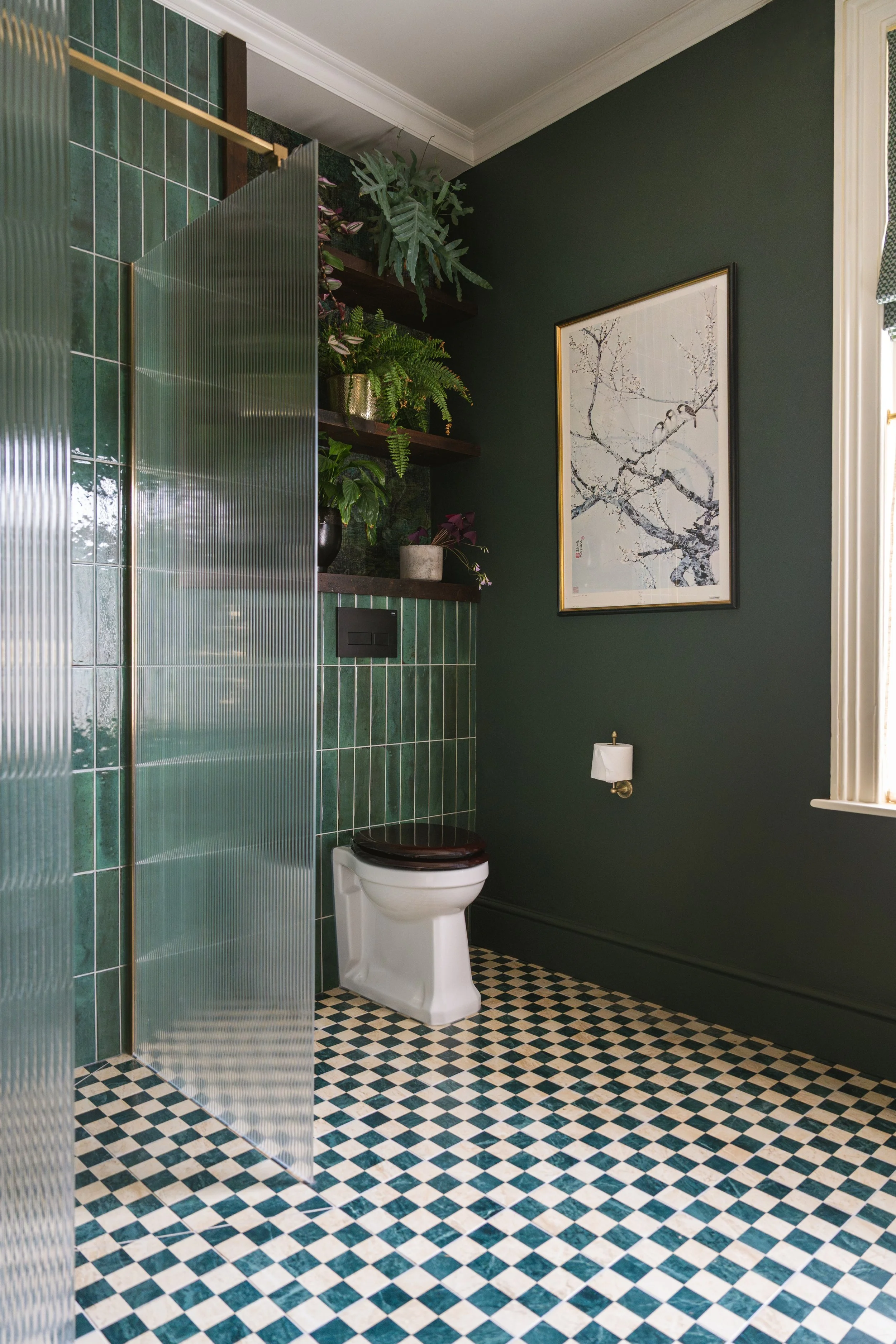 A bathroom with green tiled walls and a checkered black and white floor. There is a white toilet with a dark wood seat, a paper holder with toilet paper, and framed artwork of a tree branch on the wall. Shelves above hold various potted plants, and a textured glass shower partition is visible.