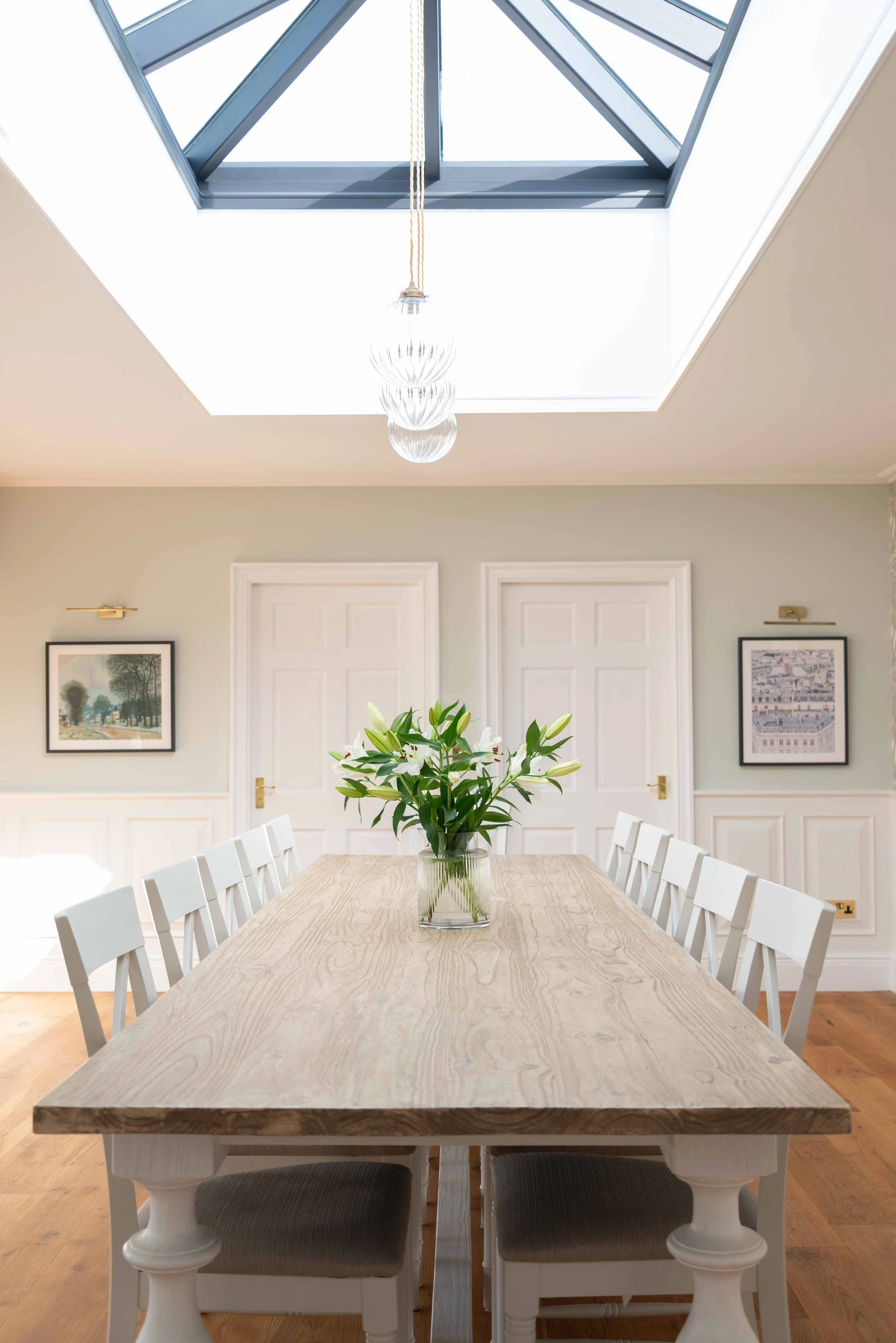 A conference room with a large wooden table surrounded by white chairs, a central vase with white lilies, framed pictures on the wall, a ceiling skylight, and a chandelier hanging from the ceiling.