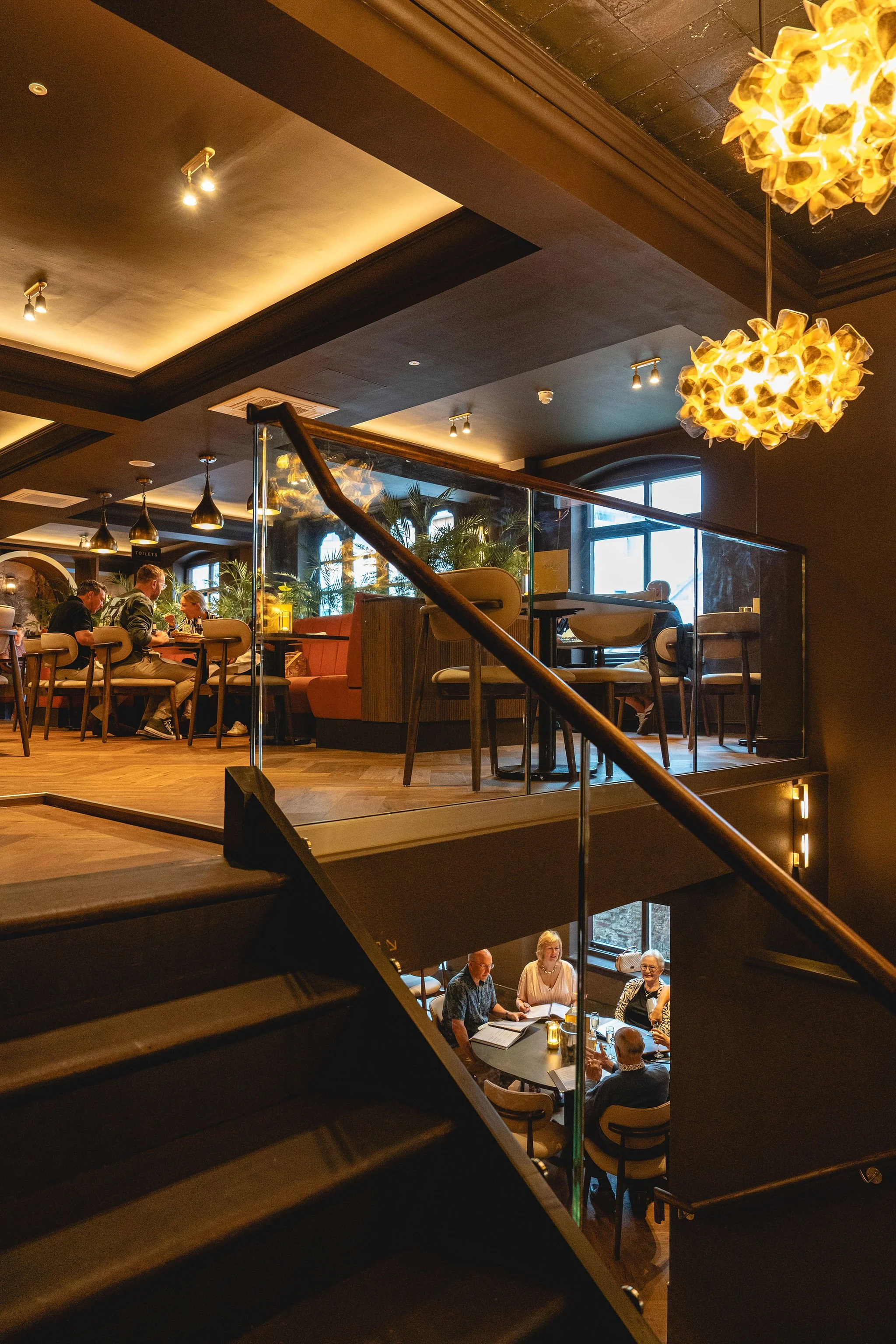 Interior of a stylish restaurant or cafe with dark walls, large windows, and modern lighting fixtures. There are multiple levels with people dining and socializing, surrounded by plants and cozy seating arrangements.