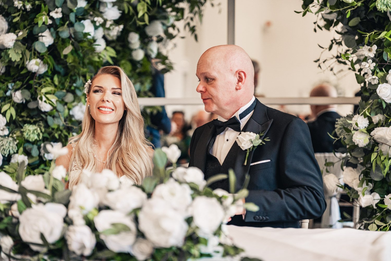A bride with long blonde hair smiling and a groom in a black tuxedo with a bow tie standing at a wedding reception, surrounded by lush green and white floral decorations.