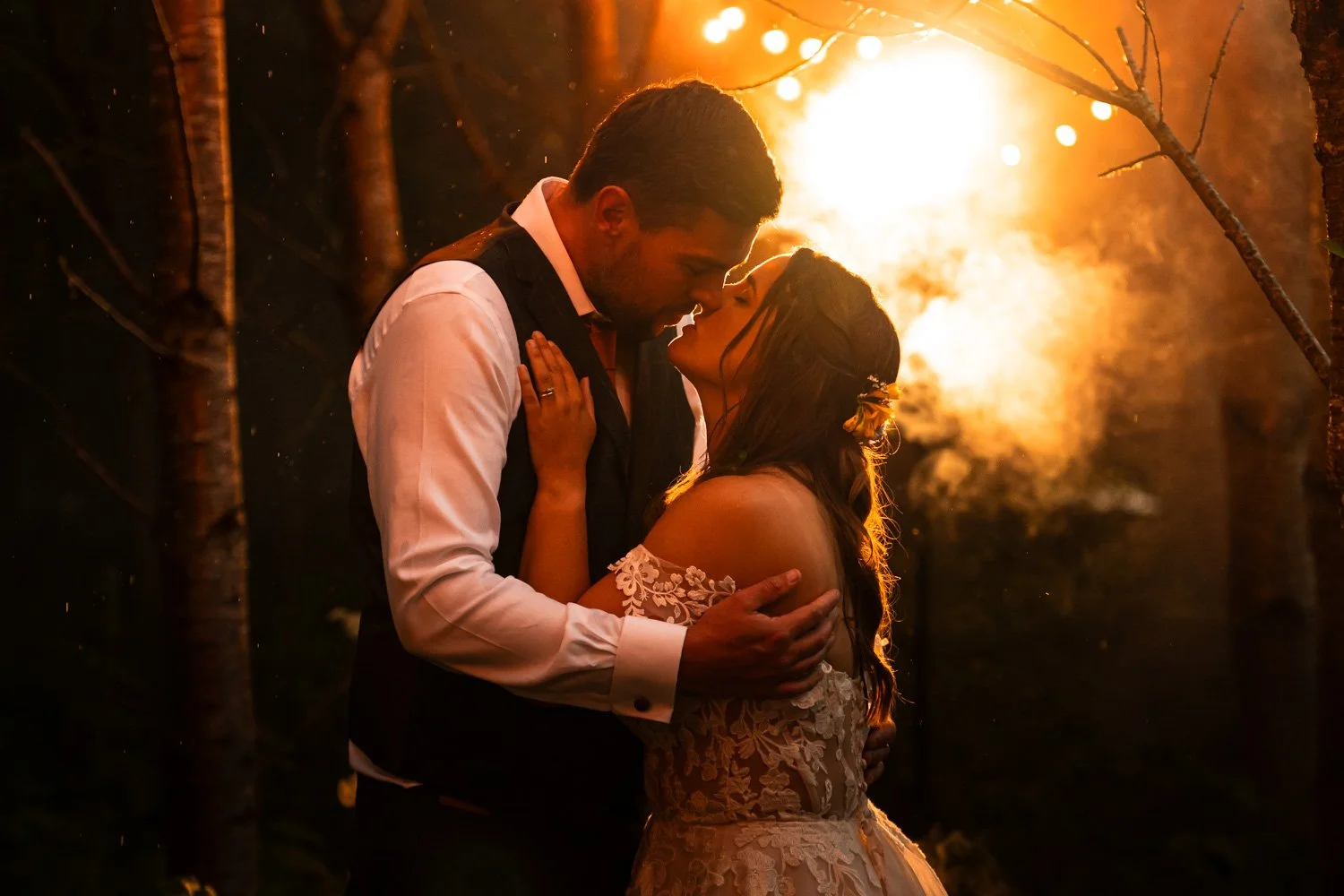 A romantic couple sharing a kiss at sunset surrounded by trees, with warm orange lighting and glowing lights in the background.