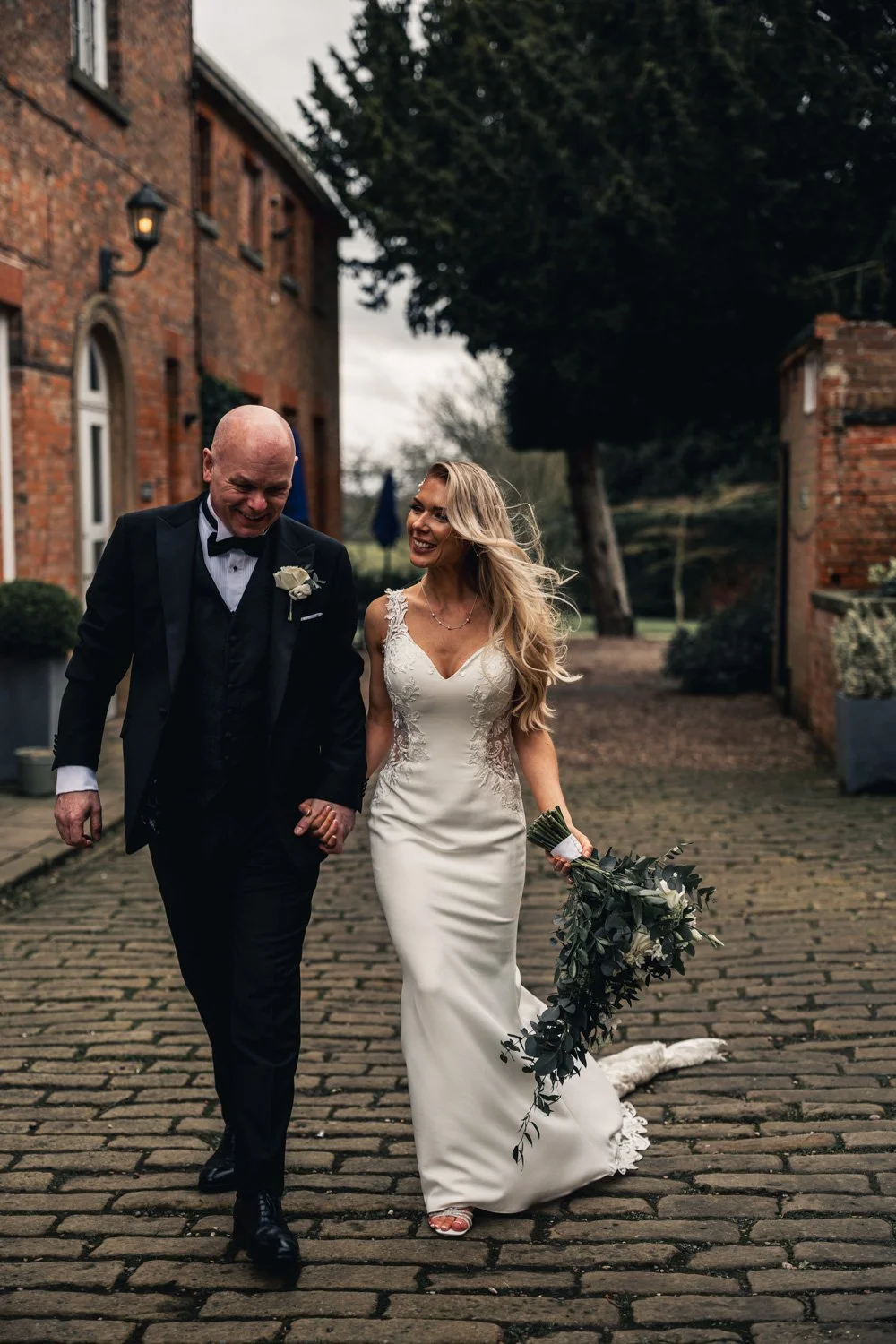 A bride in a white wedding dress holding a bouquet of greenery, walking arm-in-arm with a groom in a black tuxedo and bowtie, outside on a cobblestone path near brick buildings and trees, smiling and laughing.