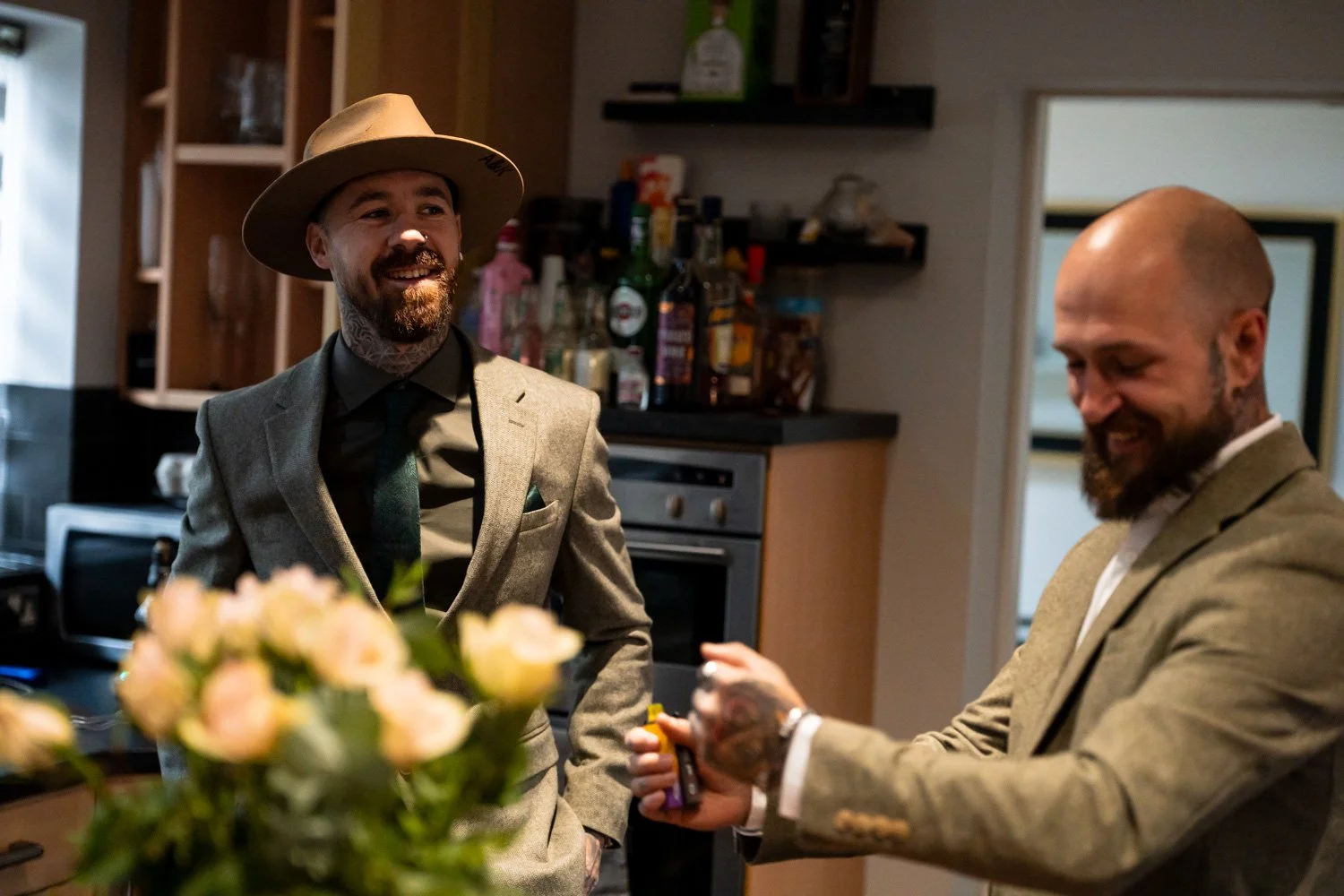 Two men with beards and tattoos dressed in suits in a kitchen, one wearing a hat, smiling, and the other holding a small bottle and smiling. There is a blurred bouquet of flowers in the foreground.