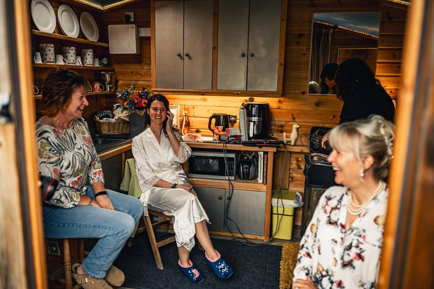 Four women inside a cozy wooden kitchen, smiling and chatting, with one in a silk robe, likely getting ready for an event, surrounded by kitchen appliances, dishes, and a mirror.