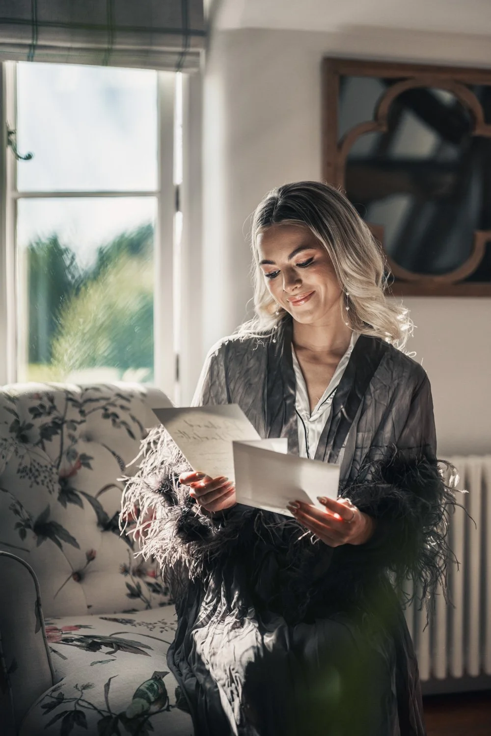 A woman sitting on a floral-patterned couch, reading handwritten letters, with sunlight coming through a window behind her.