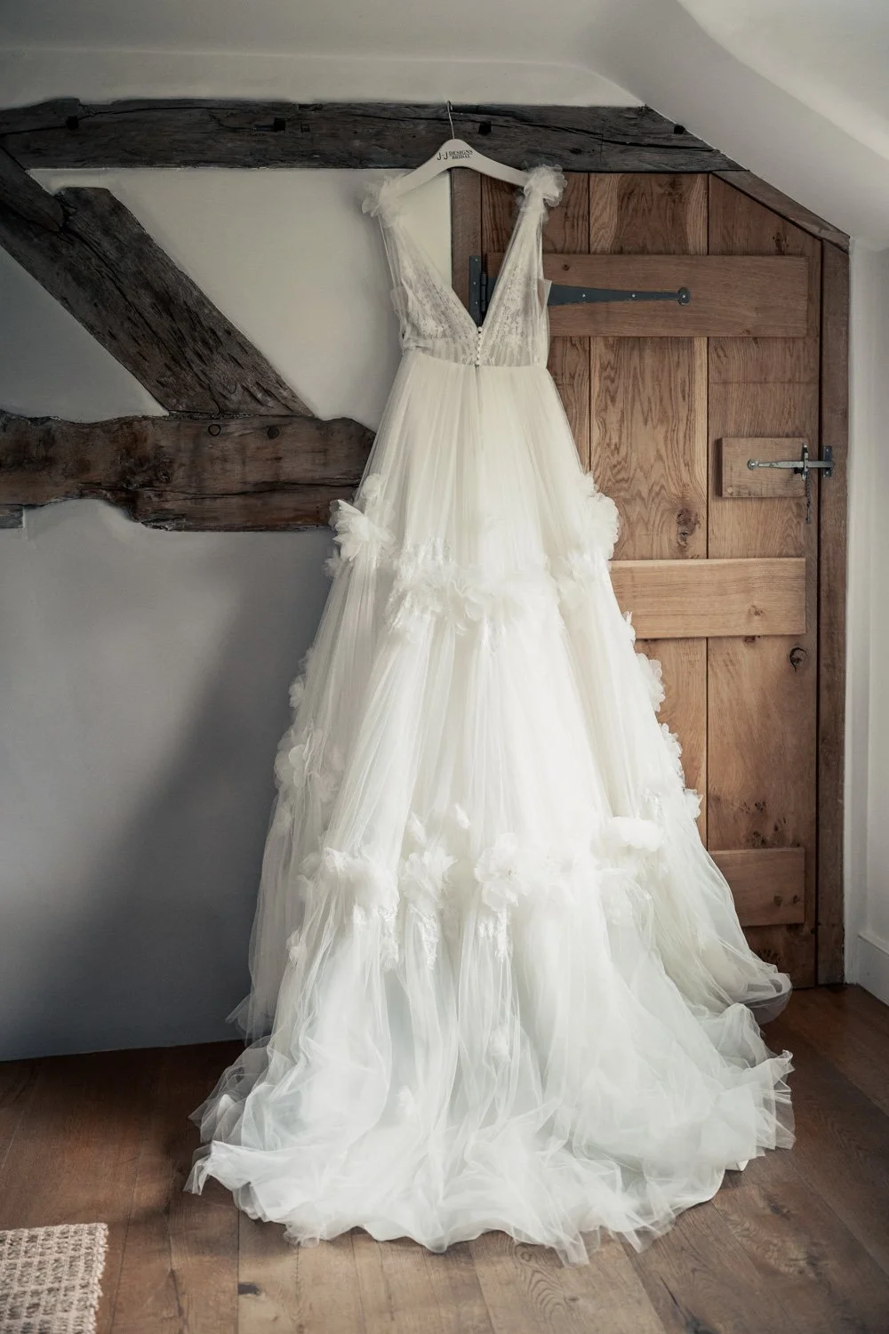 A white wedding dress with ruffles hanging on a wooden hanger on a rustic wooden barn door in a room with wooden floor and exposed dark wooden beams.