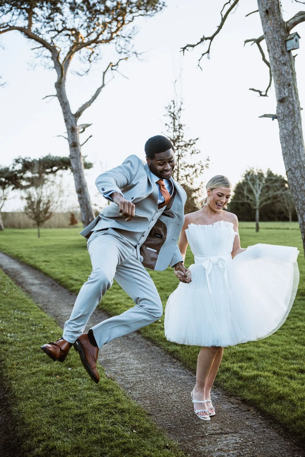A newlywed couple, the groom in a light gray suit and the bride in a white dress, holding hands and dancing joyfully outdoors on a grassy path with trees in the background during daylight.