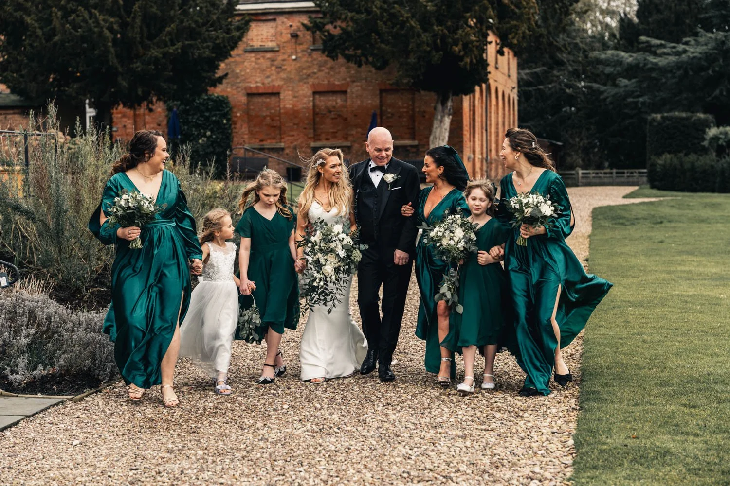A wedding party walking outdoors on a gravel path, including the bride, groom, bridesmaids, and young girls, all dressed in formal attire with the bridesmaids in teal dresses and holding bouquets, with a historic brick building in the background.