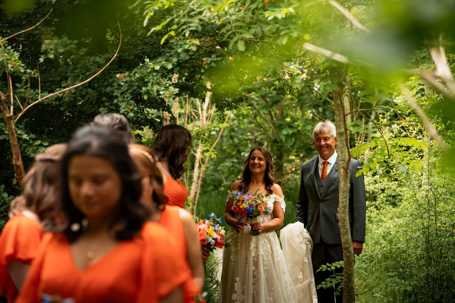 A bride and a man in a suit are smiling during a wedding ceremony in a lush green outdoor setting, with several women in orange dresses in the foreground.