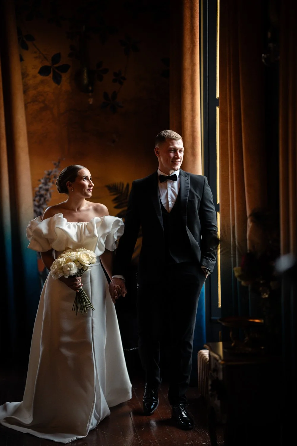 A bride and groom standing hand in hand near a large window with golden curtains in a dimly lit room, the bride holding a bouquet of white roses while looking at the groom, who is smiling and dressed in a black tuxedo with a bow tie.