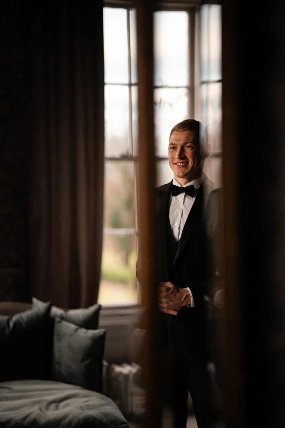 A young man in a tuxedo with a bow tie smiling and looking through a window with curtains, reflected in the glass.