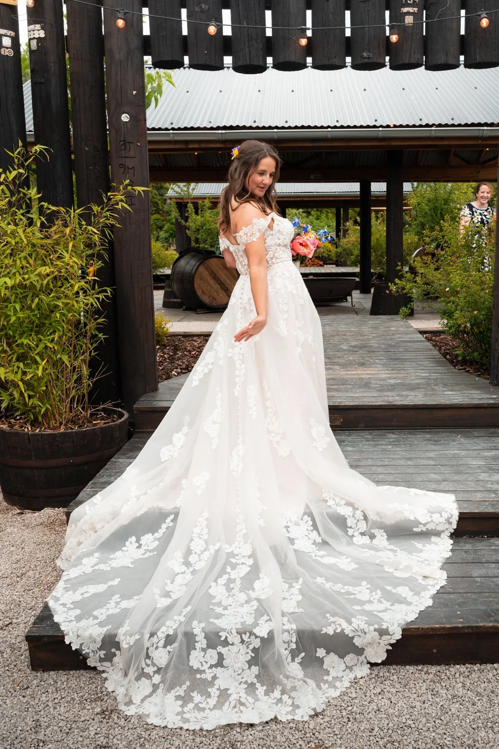 A bride in a white lace wedding gown with a long train, standing on a wooden pathway outdoors, holding a bouquet of pink and purple flowers, smiling.