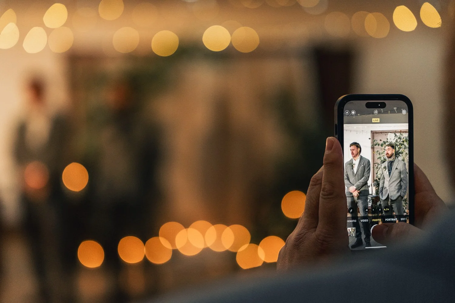 Person taking a photo of two men in suits on a smartphone at an indoor event with warm lighting and blurred background.