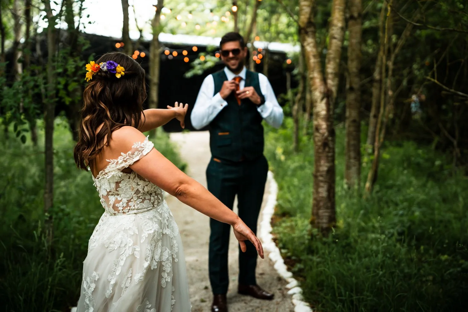 A woman in a wedding dress with floral headpiece dancing in a wooded outdoor setting, with a man in a vest and tie smiling in the background.