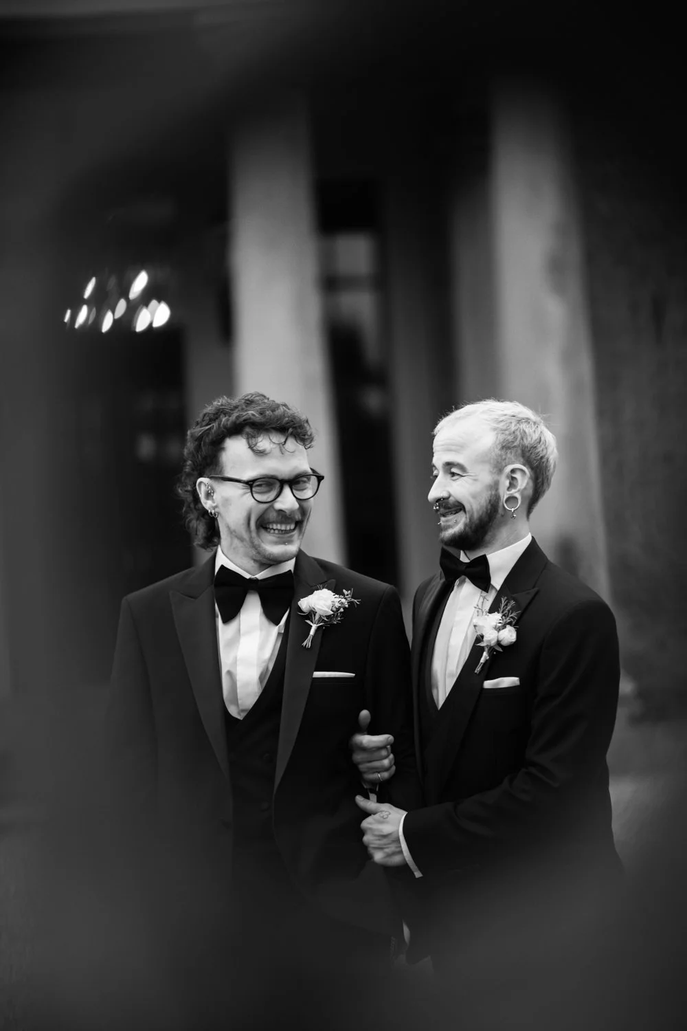 Two men in tuxedos with boutonnieres, smiling and talking at a wedding or formal event, black and white photo.