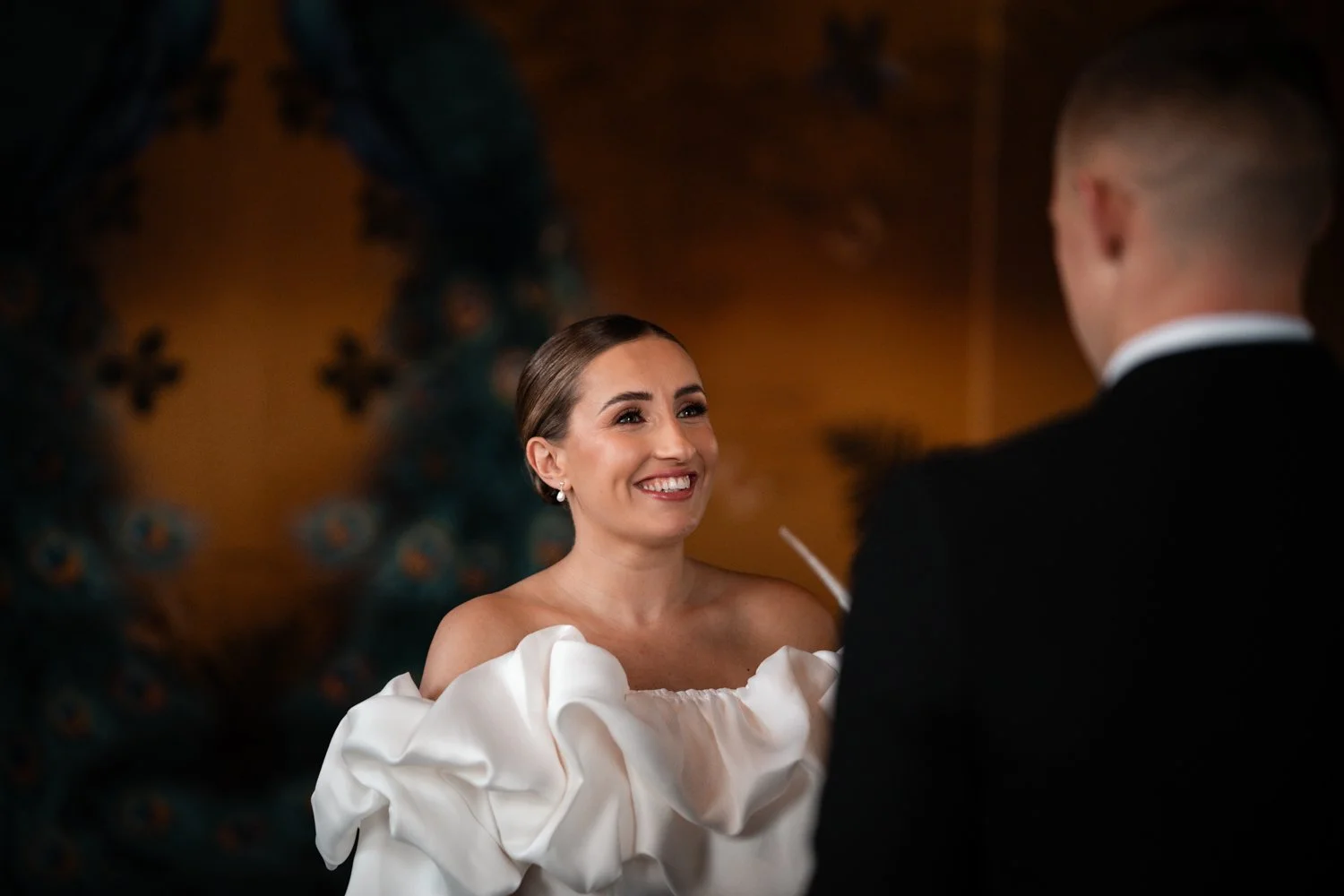 A bride in a white off-shoulder dress smiling at a groom during their wedding ceremony.