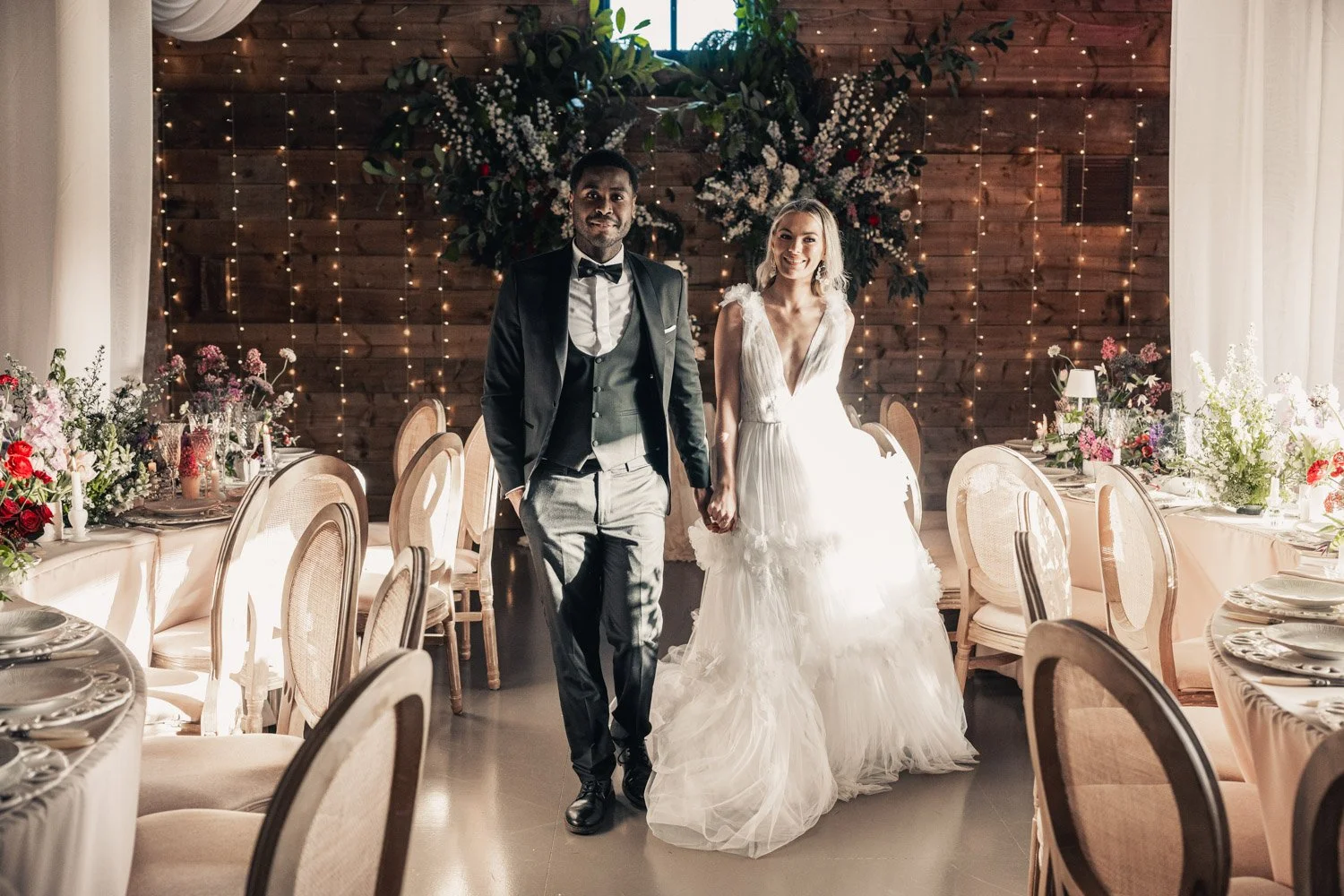 A newlywed couple walking hand in hand in a decorated wedding reception hall, with flowers and fairy lights in the background.