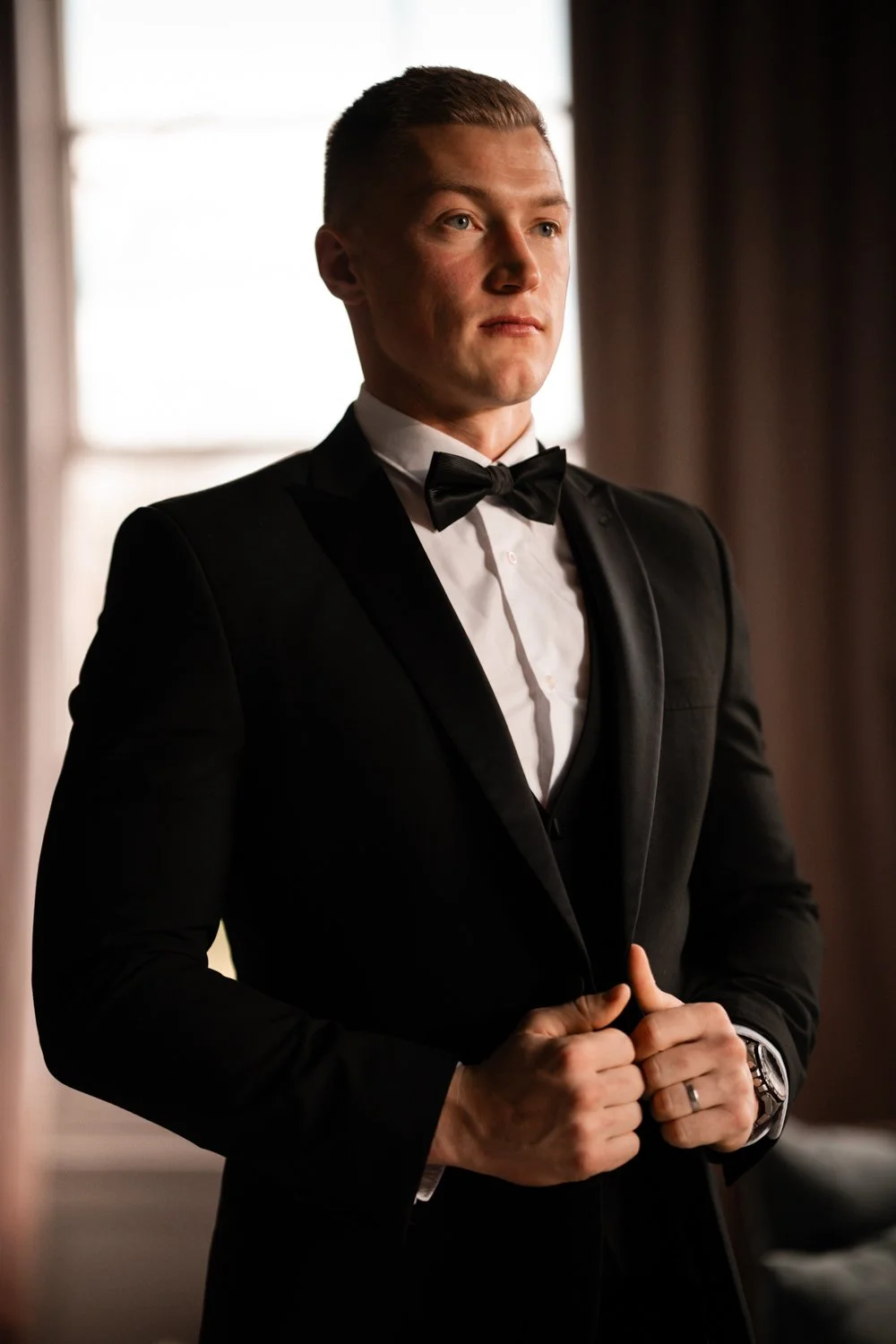 A young man in a black tuxedo with a bow tie, adjusting his jacket in front of a window with curtains.