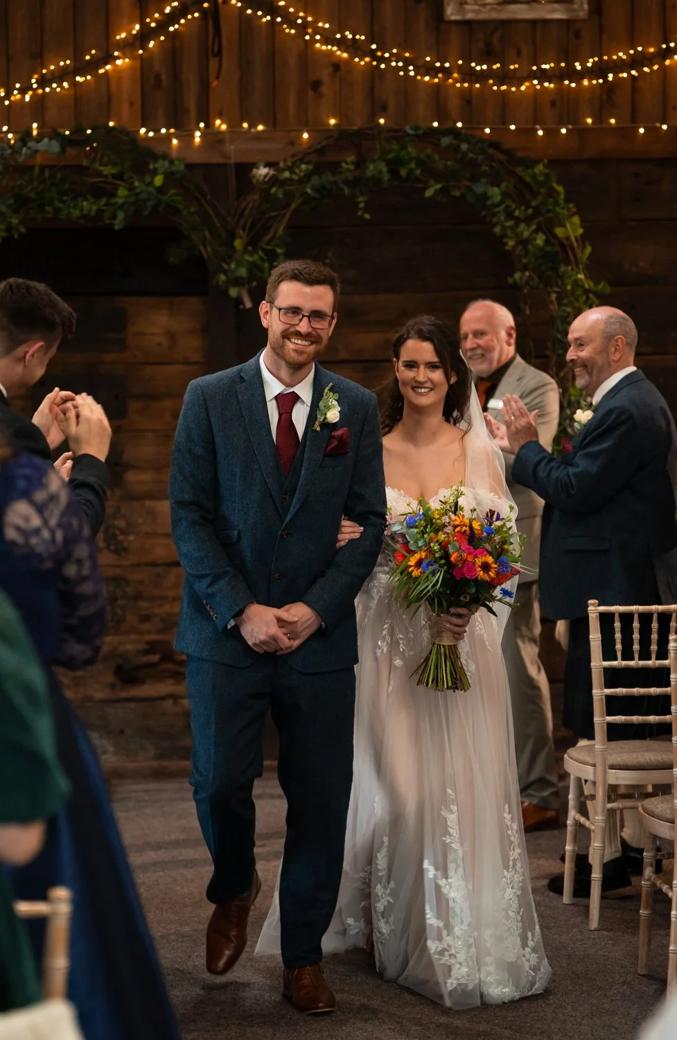Bride and groom walking down the aisle at their wedding. The bride holds a colorful bouquet and smiles, while the groom is dressed in a blue suit with a red tie and boutonnière. Guests and officiants look on and applaud, with wooden walls and string 
