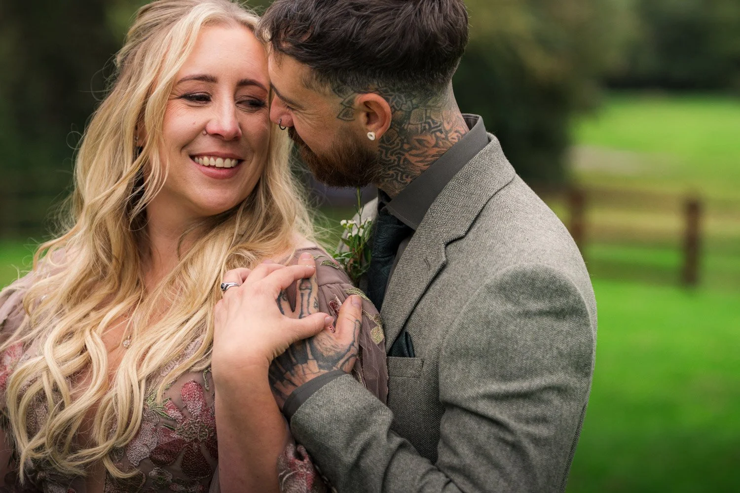 A smiling woman with long blonde hair and a man with tattoos, a beard, and short brown hair, sharing a moment outdoors. The woman is wearing a patterned dress, and the man is in a gray suit. They are close, with the woman's hand on the man's shoulder