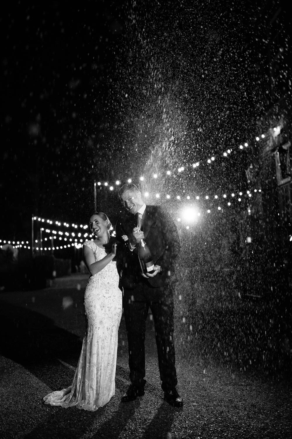 A couple dressed in formal attire celebrating with champagne at night under string lights, with confetti falling around them.