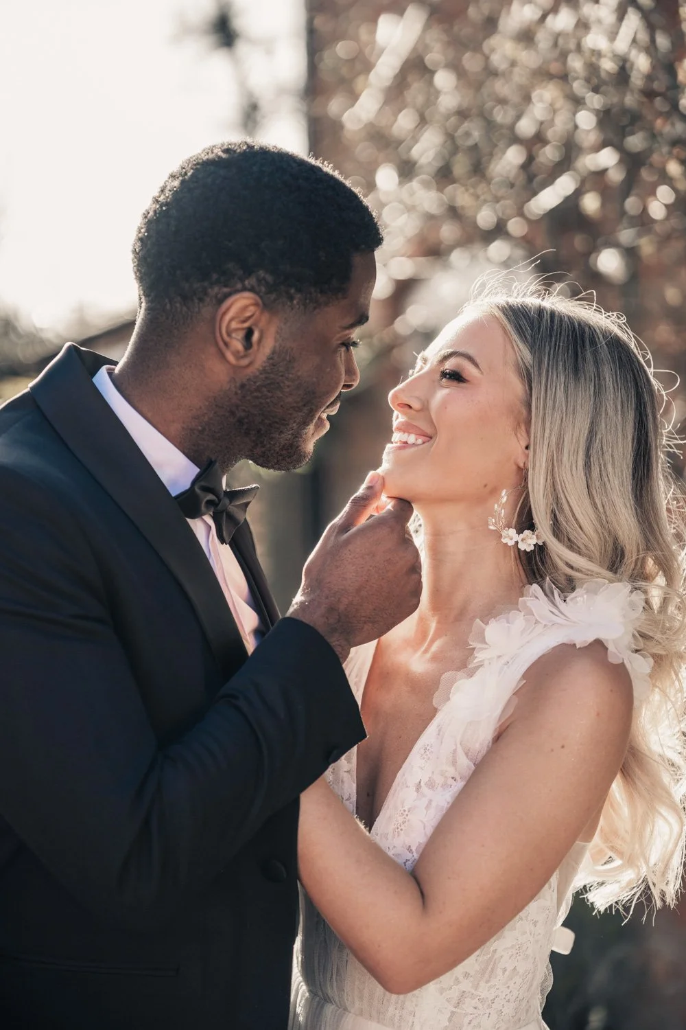 A groom in a black tuxedo gently touches a bride's chin as they look into each other's eyes, outdoors with blurred trees and sunlight in the background.