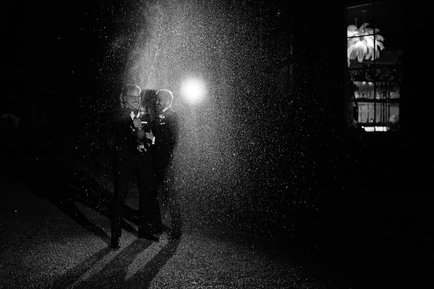 Three men in tuxedos standing in a wet street at night, illuminated from behind, with rain or spray falling around them.