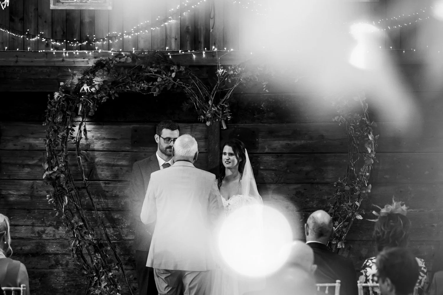 Black and white photo of a wedding ceremony with a bride and groom standing under an arch decorated with leaves and flowers, facing an officiant. Guests are seated, watching the ceremony in a rustic indoor setting with wood-paneled walls, string ligh