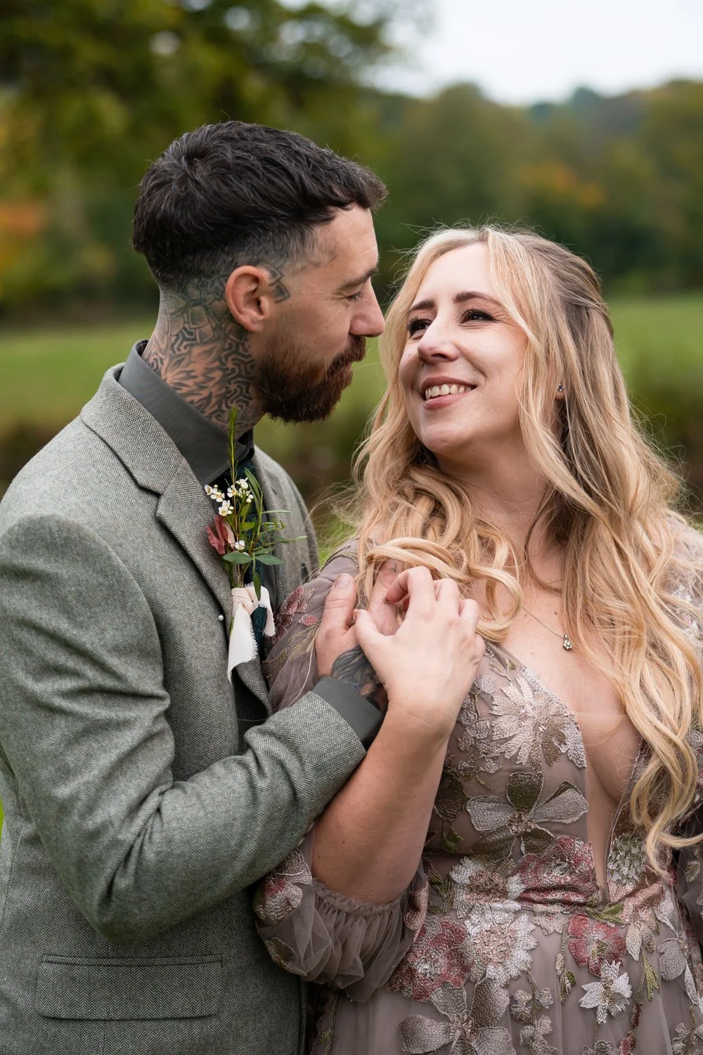 A couple facing each other outdoors, smiling and holding hands, with a scenic background of trees and greenery.